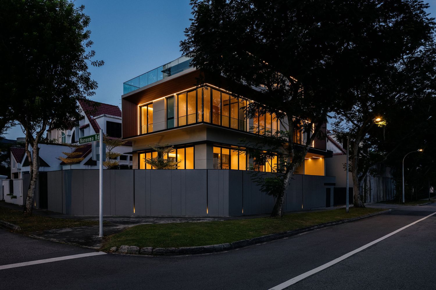 Modern two-story corner house with glowing warm windows and a glass-railed rooftop terrace, seen from the street at dusk.