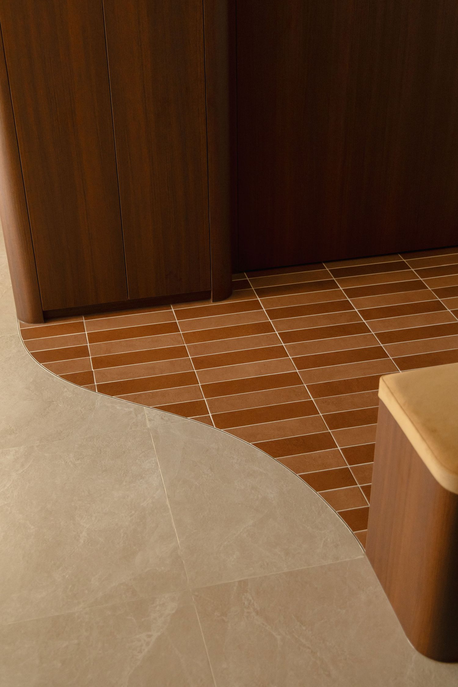 Curved transition between gray stone floor tiles and terracotta brick-pattern tiles beside warm wood cabinetry and a rounded wooden bench.