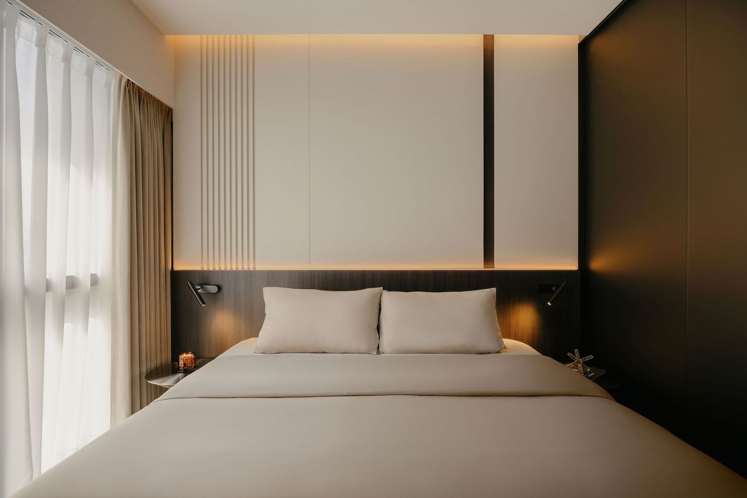 Modern minimalist bedroom with a neutral bed, cream pillows, warm backlit paneling, dark wardrobe and sheer curtains.