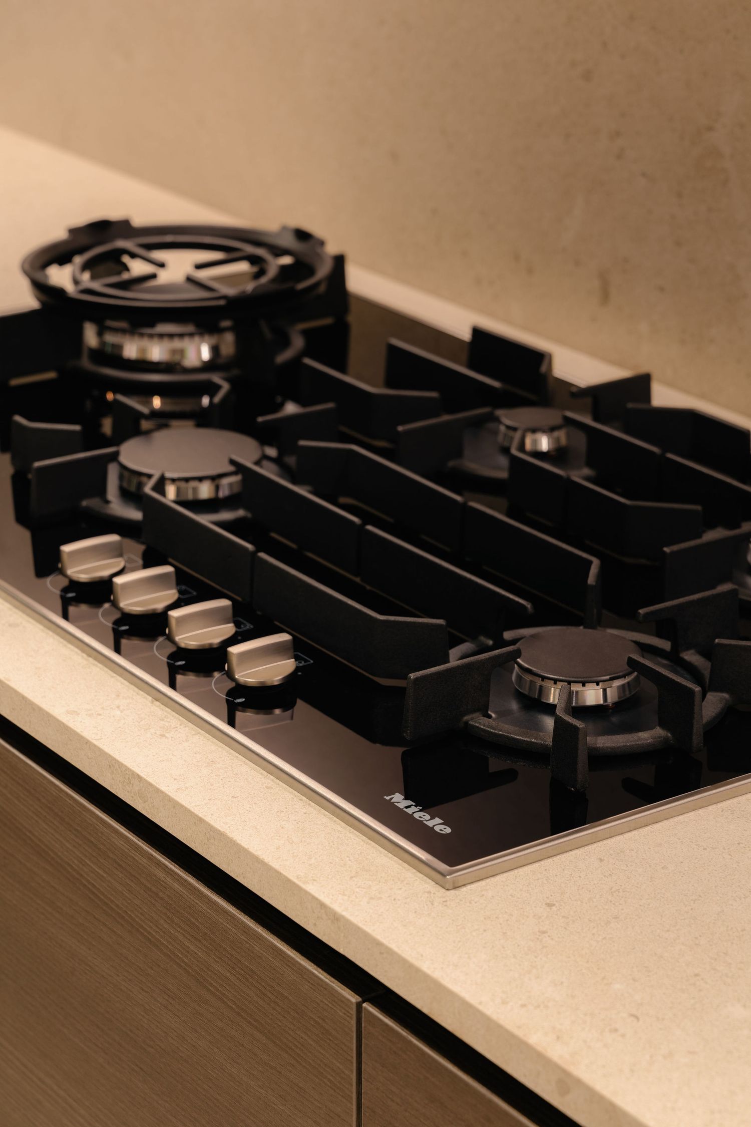 Miele black glass gas cooktop with four burners, cast iron pan supports and chrome control knobs set into a light stone countertop.