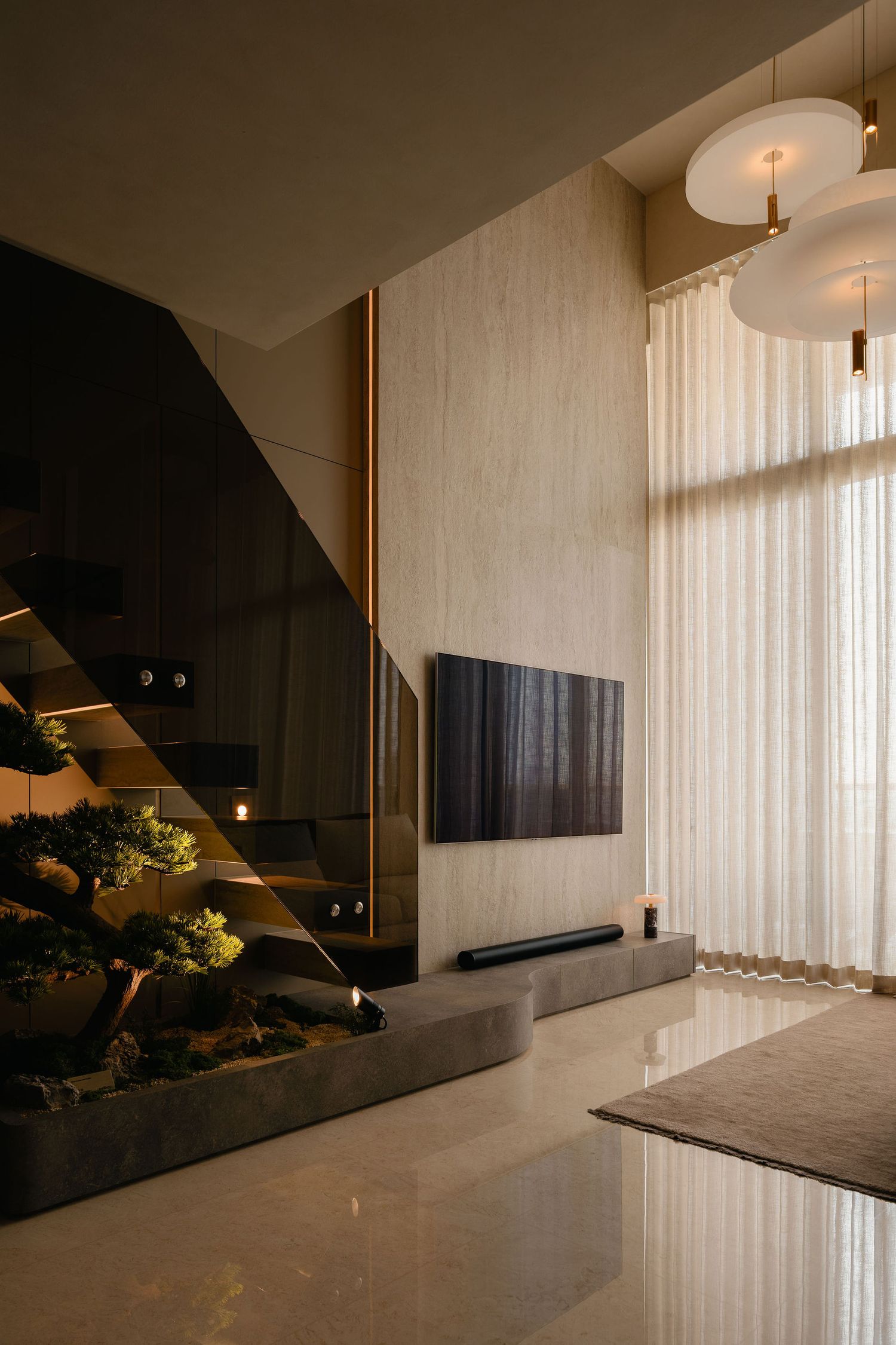 Modern double-height living room with a wall-mounted TV, floating staircase, indoor bonsai planter, sheer curtains and pendant lights.