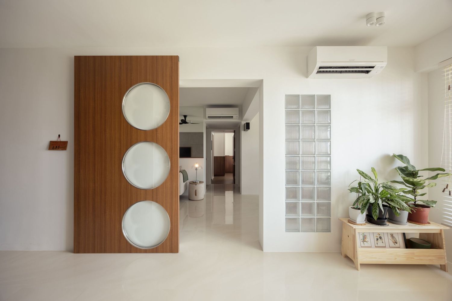 Modern apartment entryway featuring a wooden sliding door with circular windows, glass block wall, and tropical plants on a wooden shelf.