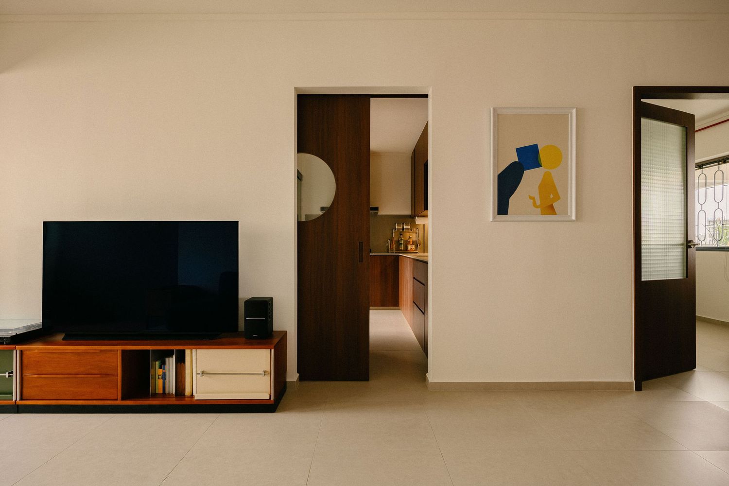 Minimalist living room with a flat-screen TV on a wooden media console, abstract wall art, and an open doorway leading to a kitchen.