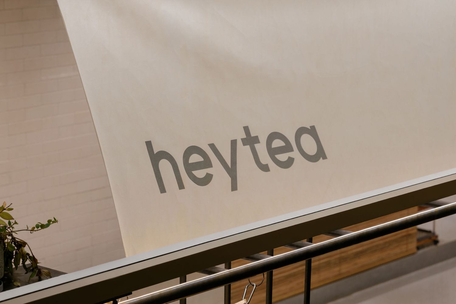 Heytea logo in gray lettering on a cream wall above a wooden ledge and black metal railing inside a modern tea shop.