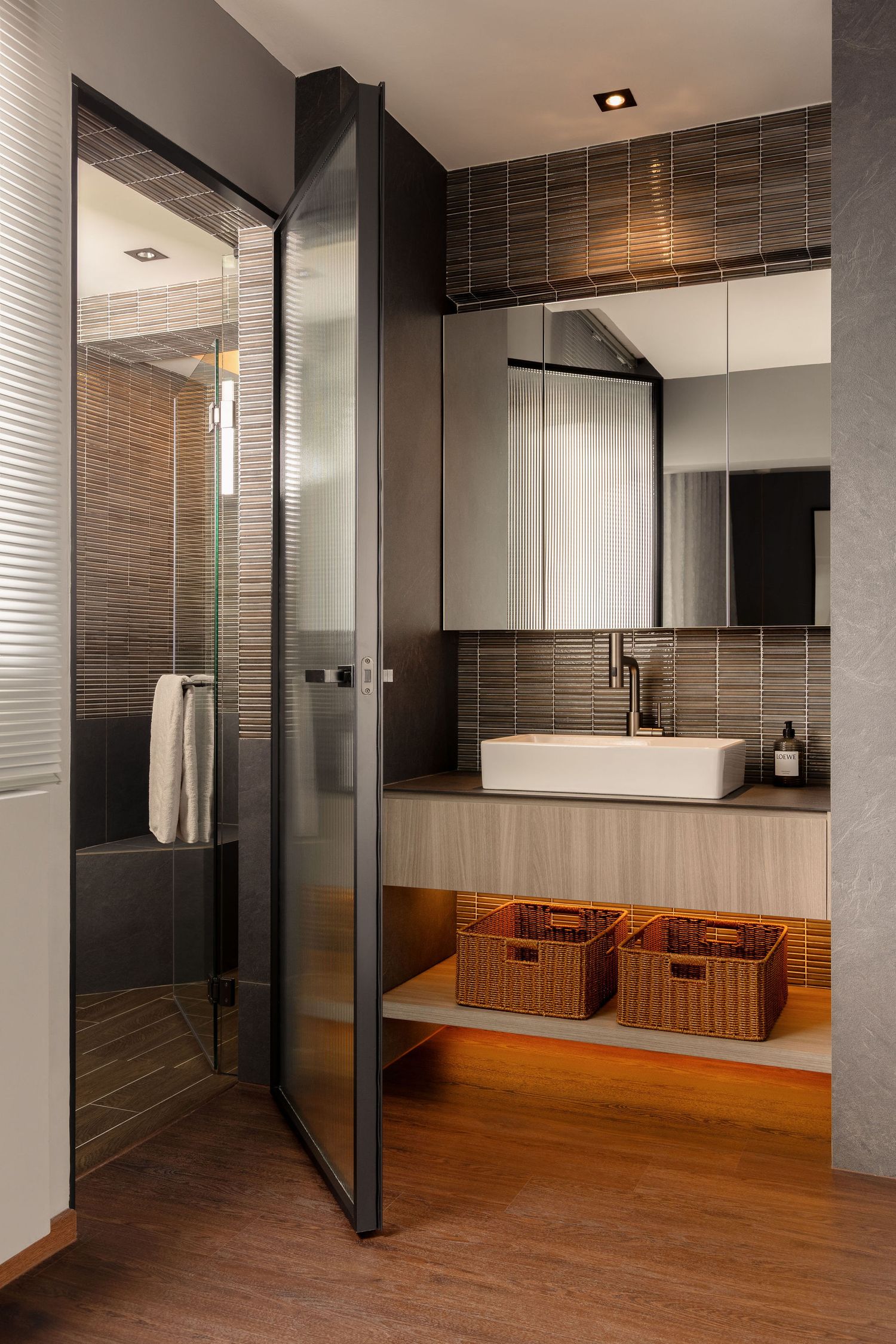 Modern bathroom with glass shower door, vessel sink, wood vanity with wicker baskets, and dark tile backsplash.