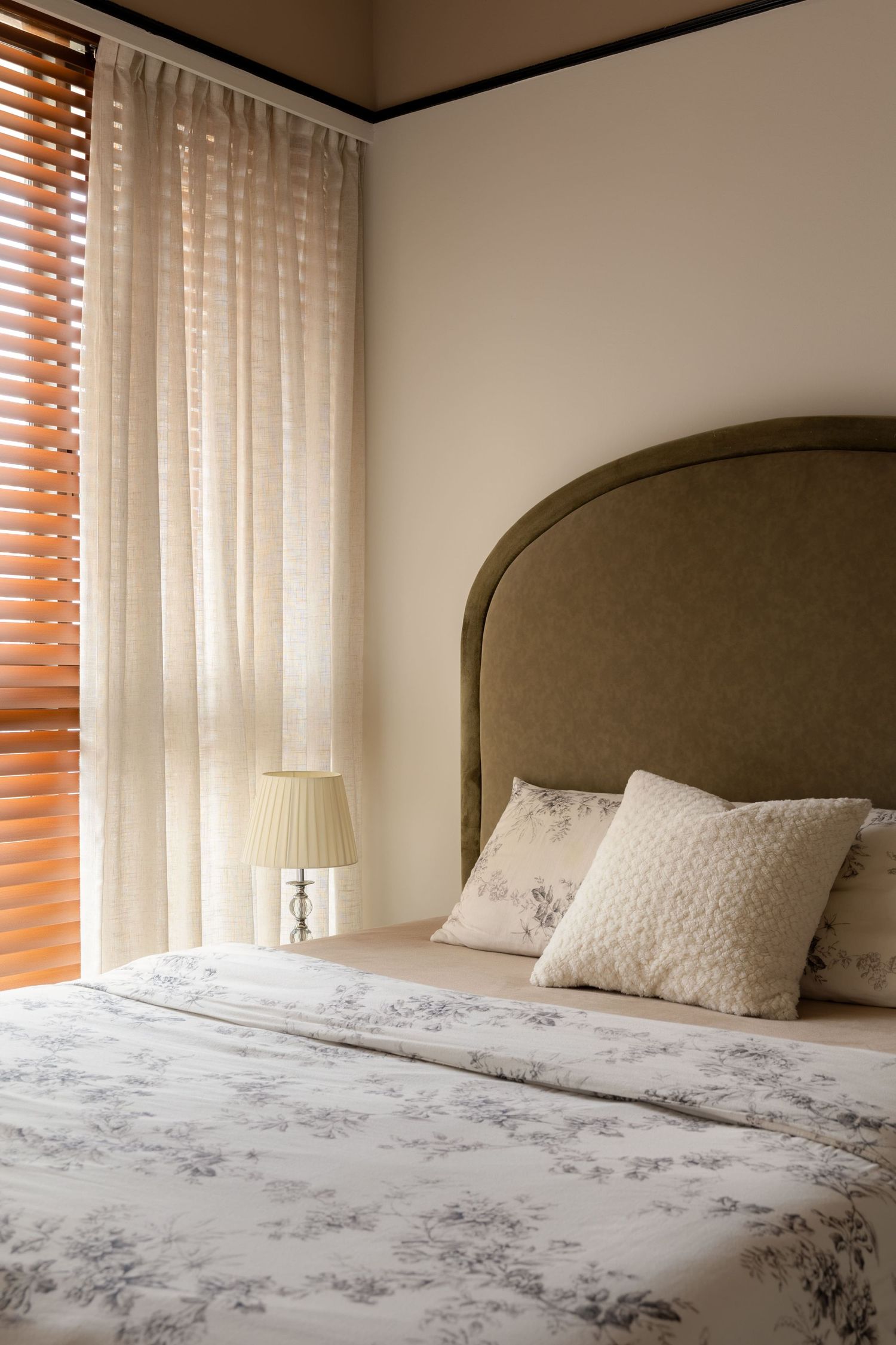 Elegant bedroom with olive green upholstered headboard, floral bedding, sheer curtains, wooden blinds, and a classic table lamp.