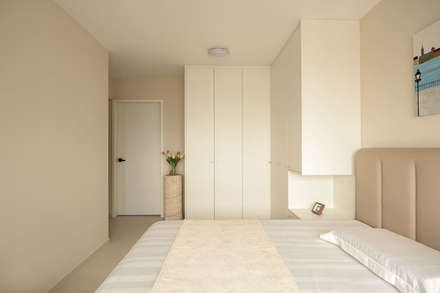 Minimalist bedroom with beige walls, built-in white wardrobes, a large bed with neutral bedding, and a small floral arrangement.