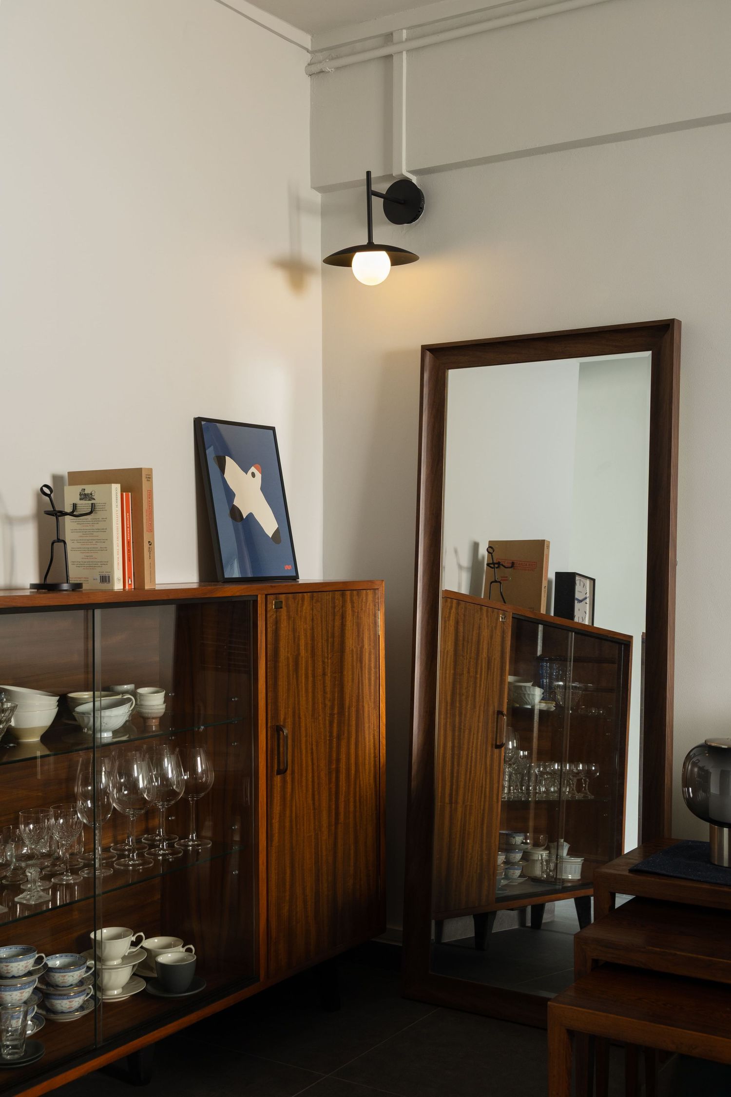 A wooden sideboard with glass display shelves holds wine glasses and dinnerware against a white wall in a dining room.