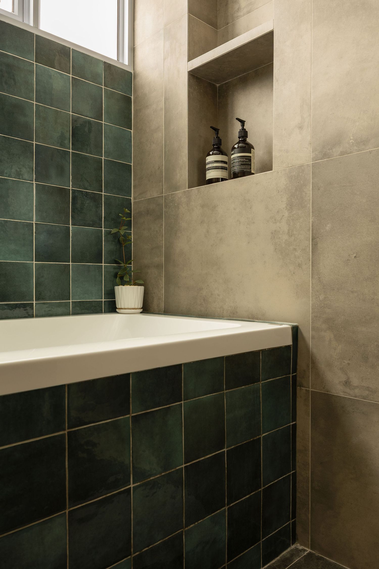 Modern bathroom with dark green square tiles surrounding a white bathtub, concrete walls, and a niche shelf with Aesop products.