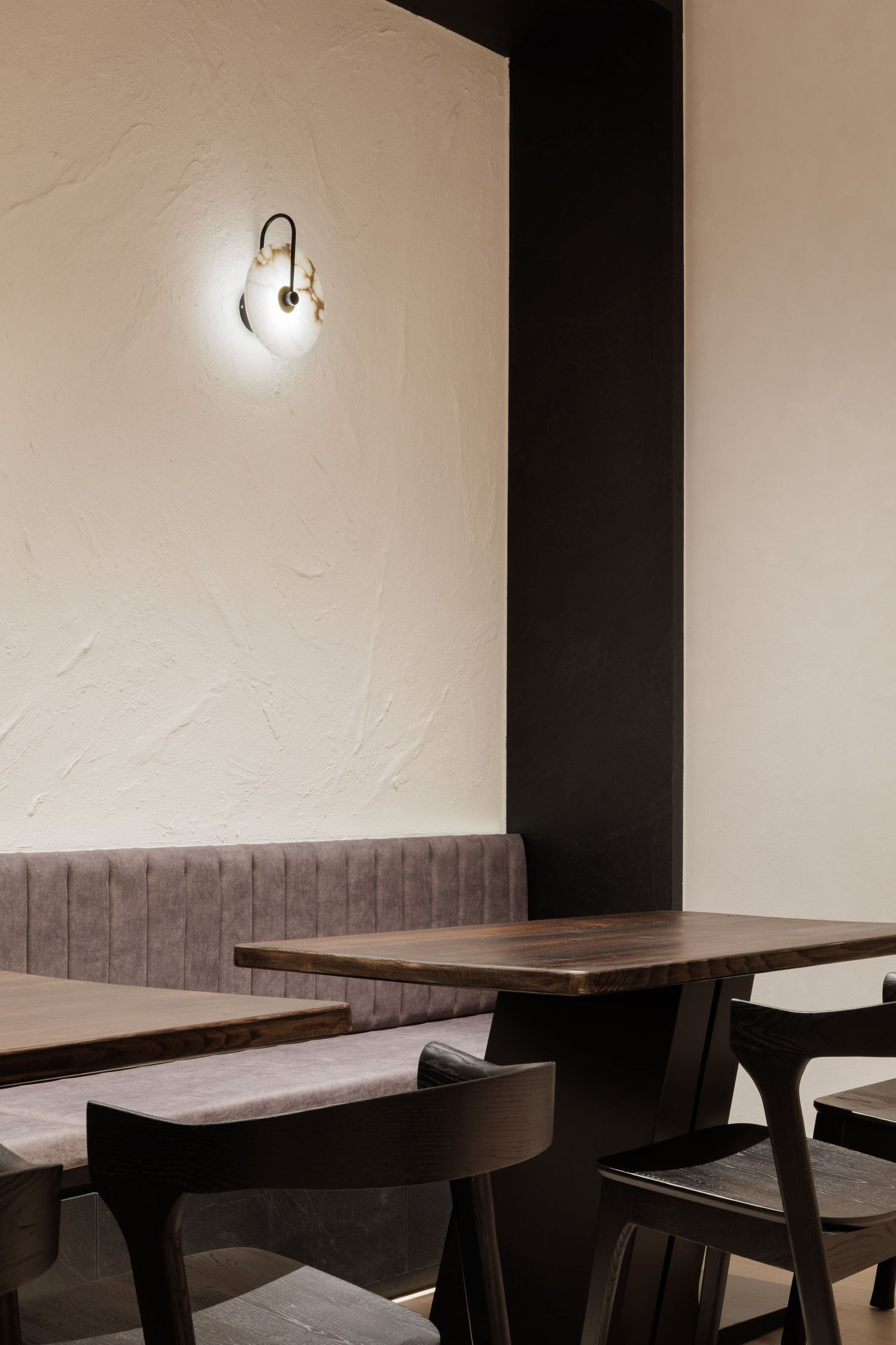 Modern restaurant interior with wooden tables, gray upholstered bench seating, and minimalist wall sconce lighting.