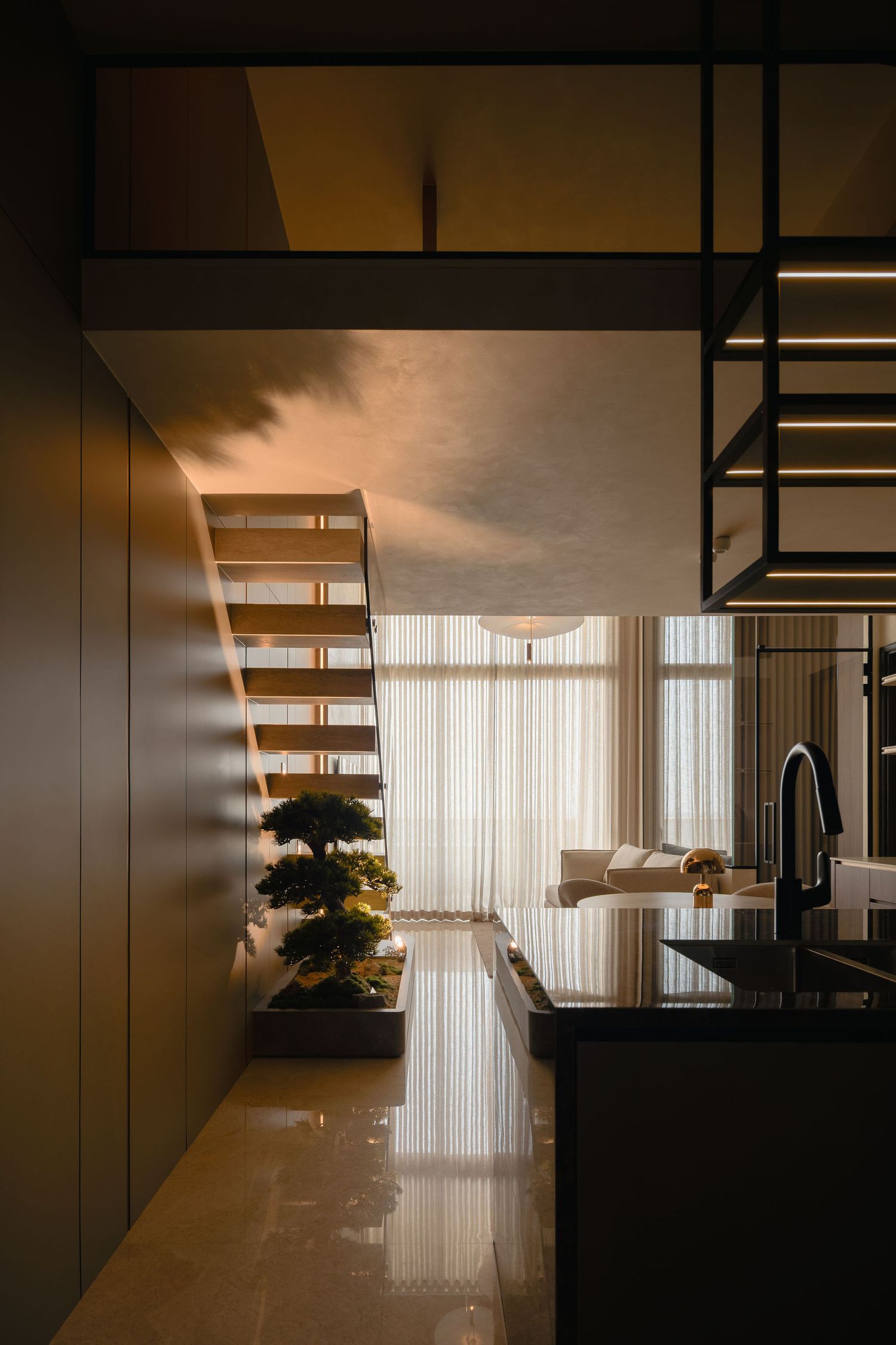 Modern loft interior with a floating wooden staircase, bonsai planter, glossy marble floor and black kitchen island under warm lighting.