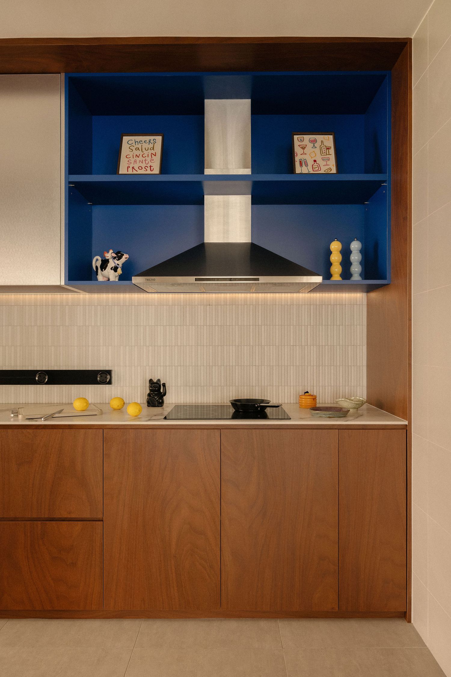 Modern kitchen with walnut wood cabinets, cobalt blue open shelving, black range hood, induction cooktop and warm under-cabinet lighting.