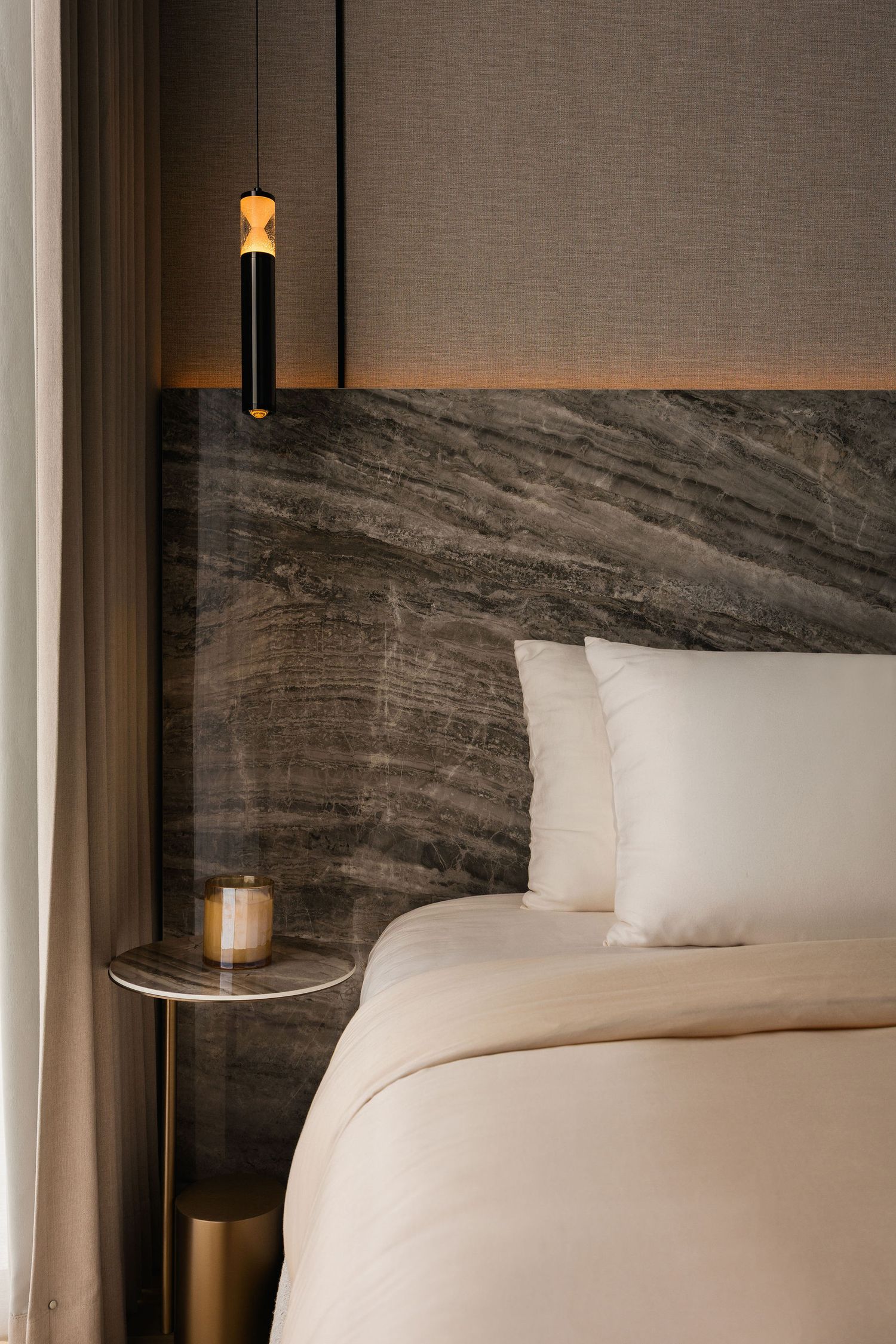 Luxury bedroom with a gray marble headboard, cream bedding, a gold pendant light and a round marble side table with a brass base.