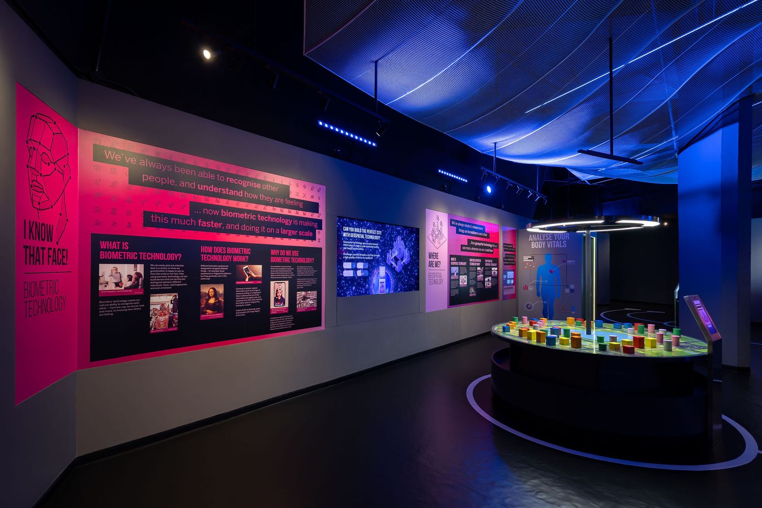 Colorful museum exhibit with pink and blue illuminated walls, interactive displays, and a round glowing installation.