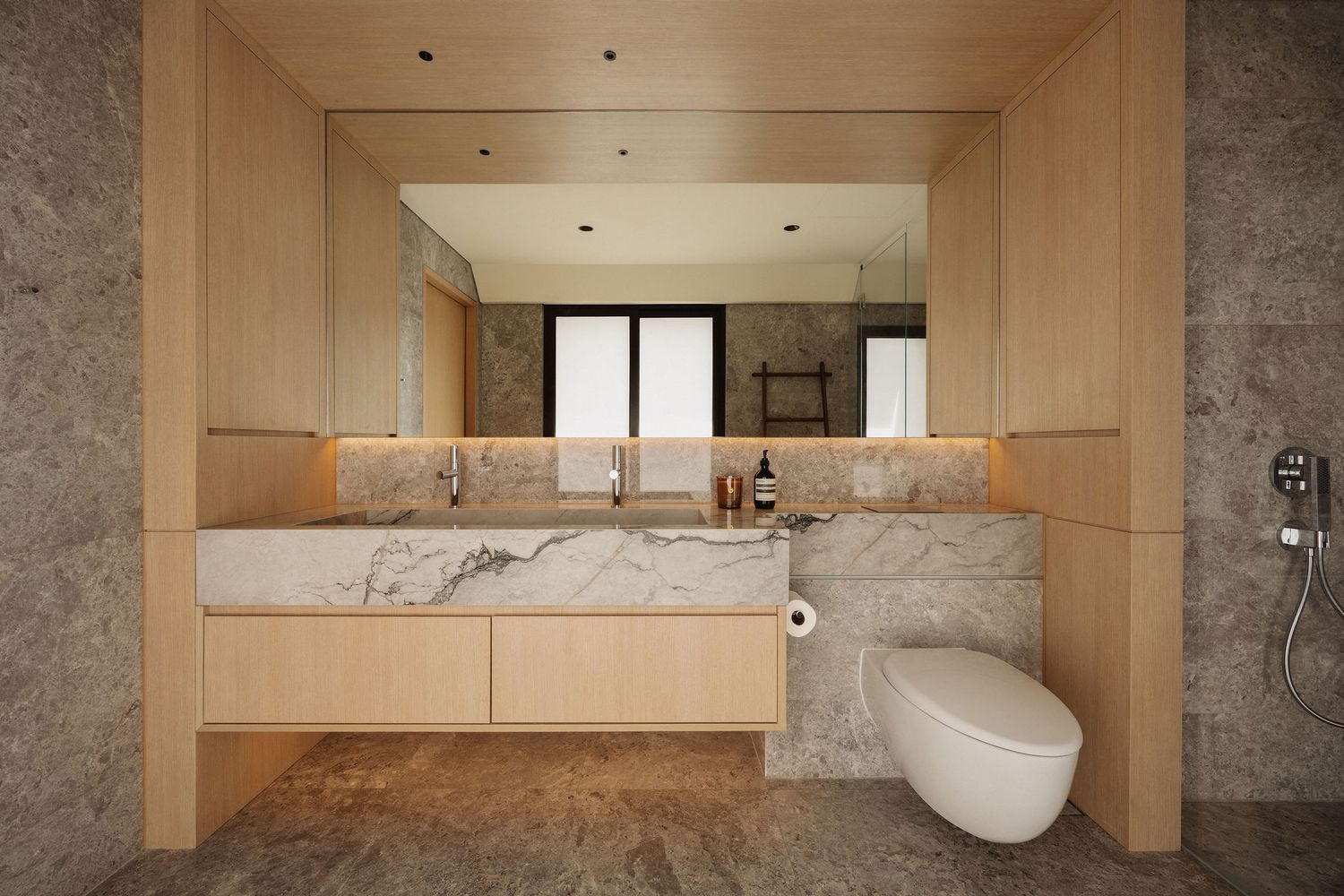 Modern bathroom with marble vanity, wood cabinetry, wall-mounted toilet, and large mirror with warm lighting.