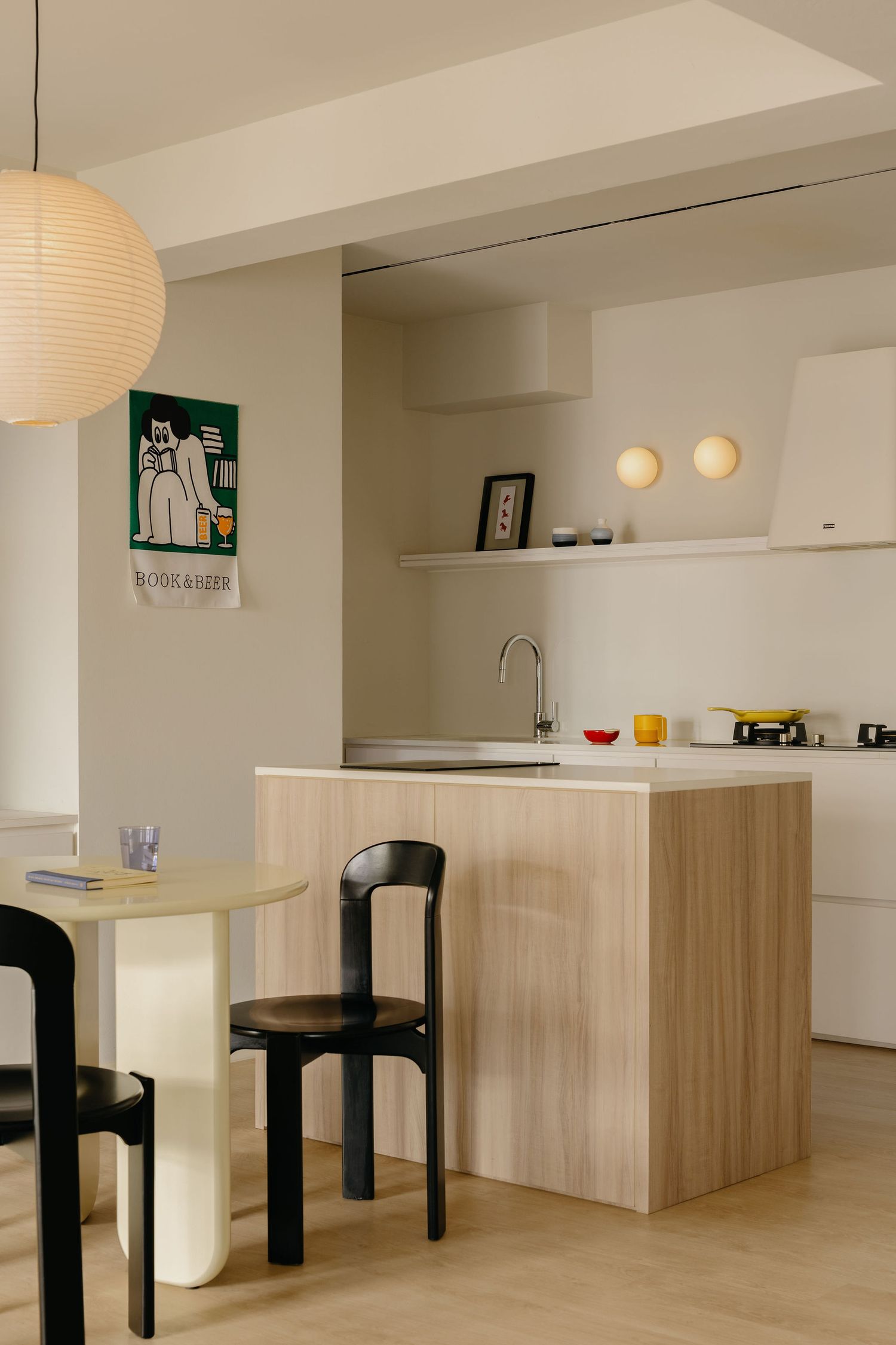 Modern Japandi kitchen with light wood island, white cabinets, black chairs, pendant lamp, and colorful art print on wall.