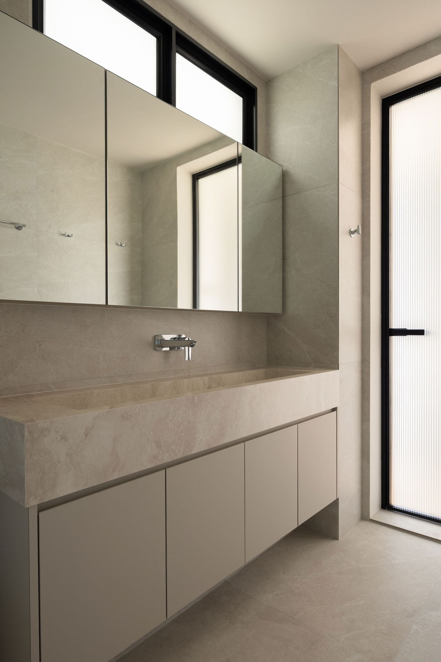 Modern minimalist bathroom with stone vanity, wall-mounted faucet, mirrored cabinets, and large windows in neutral tones.