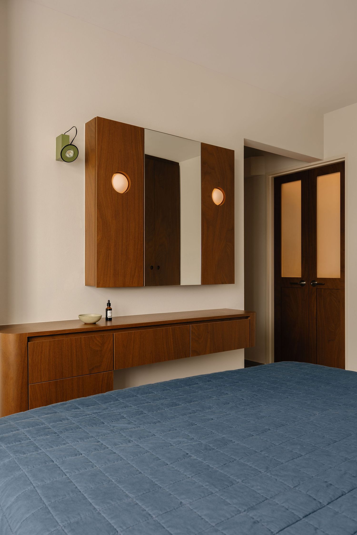 Mid-century modern bedroom with walnut floating dresser, mirror cabinet, round wall sconces, and blue quilted bedspread.