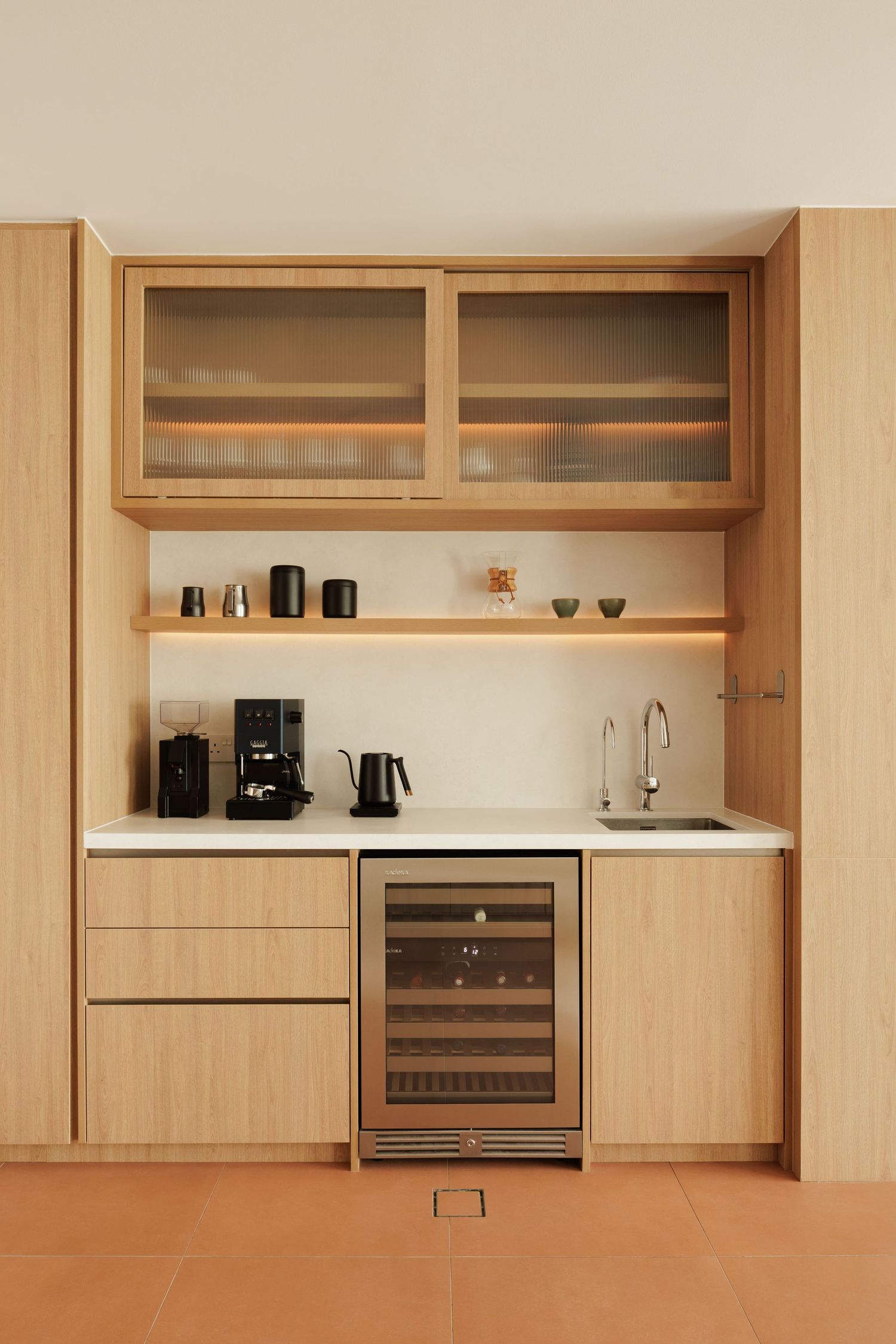 Modern minimalist coffee bar with light wood cabinets, wine fridge, and coffee maker setup on white countertop.