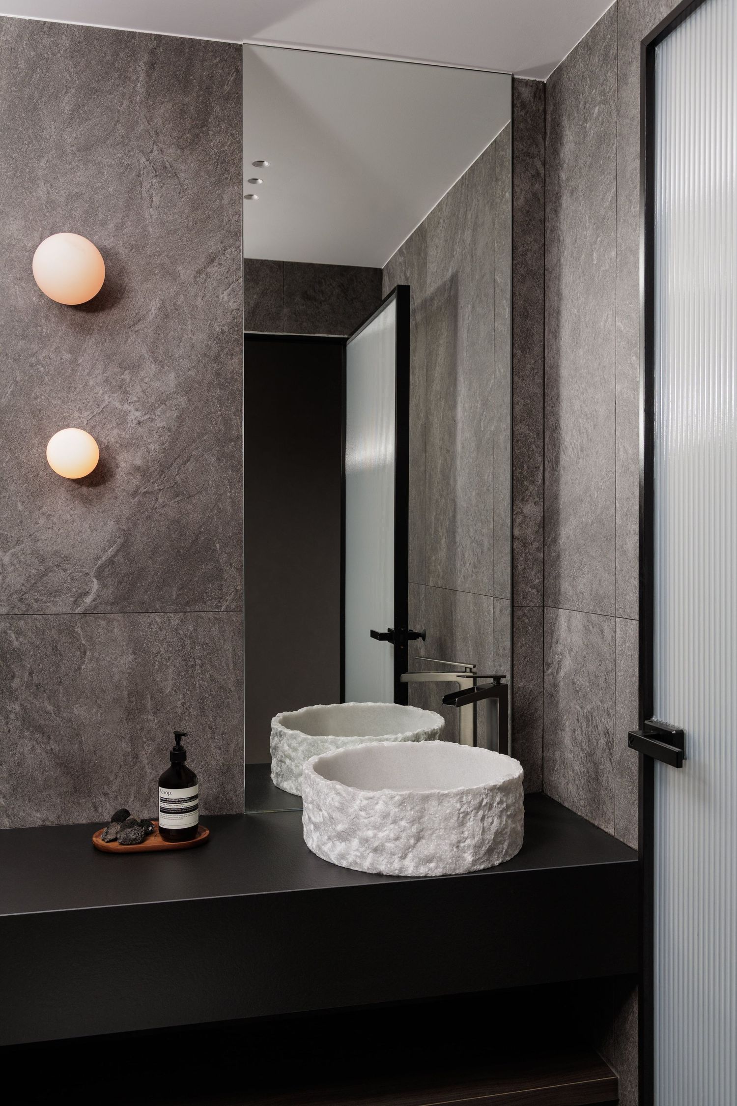 Modern bathroom interior with dark gray marble walls, black countertops, and round vessel sinks illuminated by copper sconces.