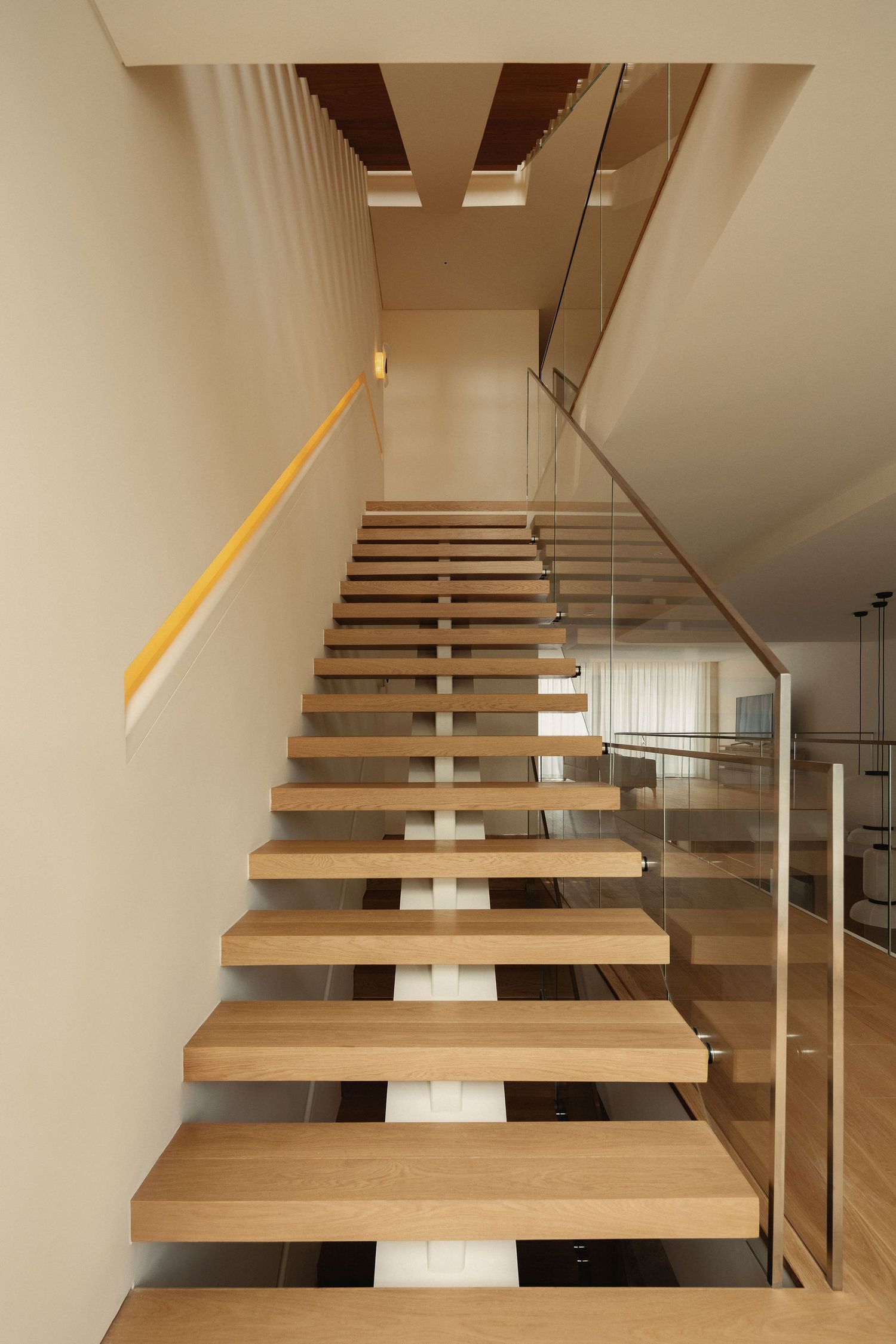 Modern floating wooden staircase with glass balustrade and yellow handrail in a contemporary open-plan home interior.