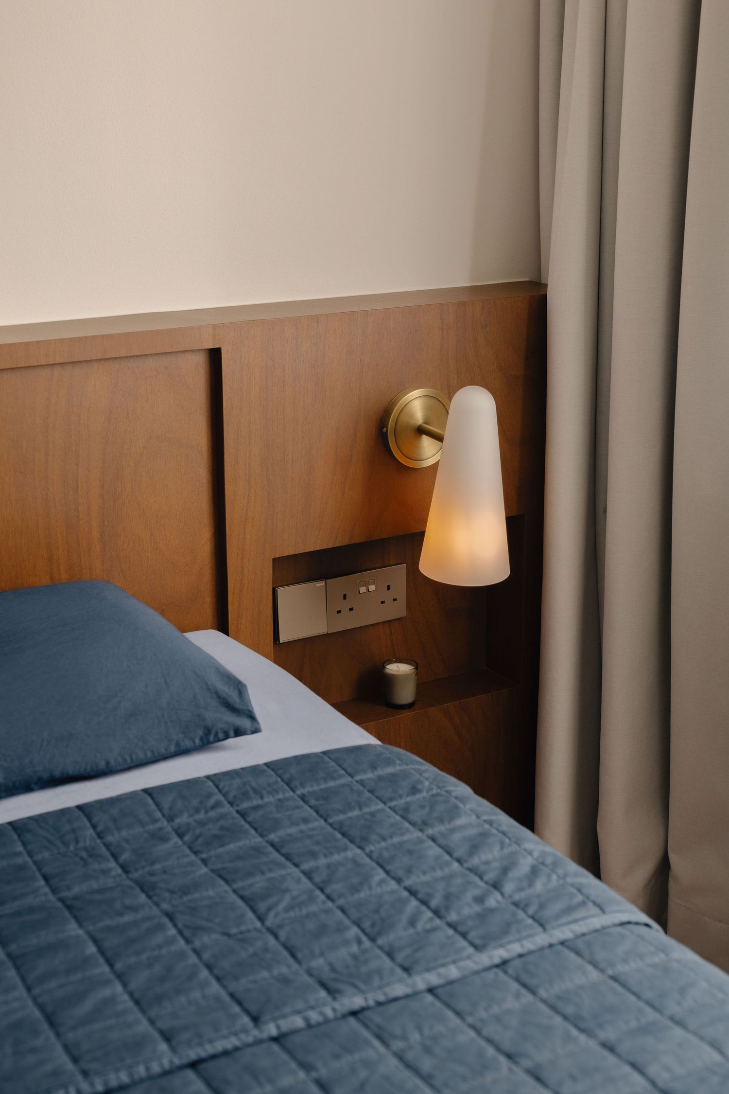 Modern hotel bedroom with teal quilted bedspread, wooden headboard, and glowing brass wall sconce nightlight.