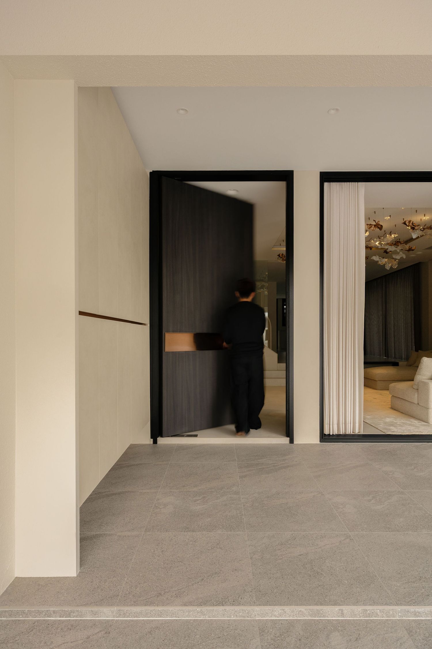 Person entering a modern interior space with dark wood paneling, black-framed doors, and elegant chandelier visible through window.
