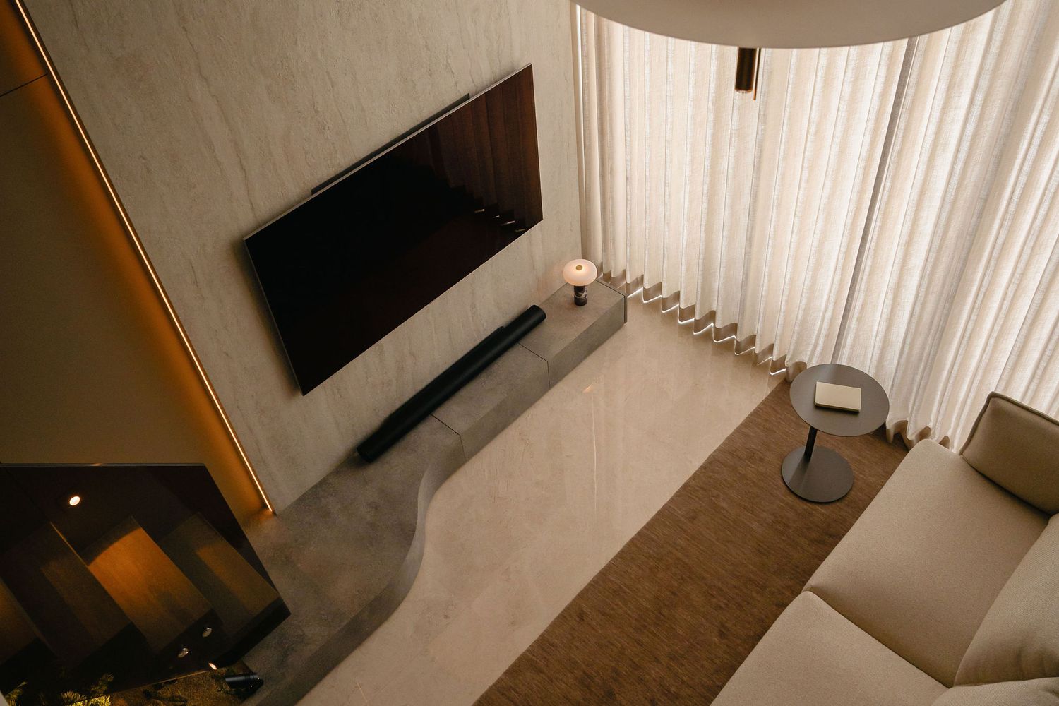 Modern living room with a wall-mounted TV, sleek soundbar on a curved console, sheer curtains, a beige sofa and round side table.