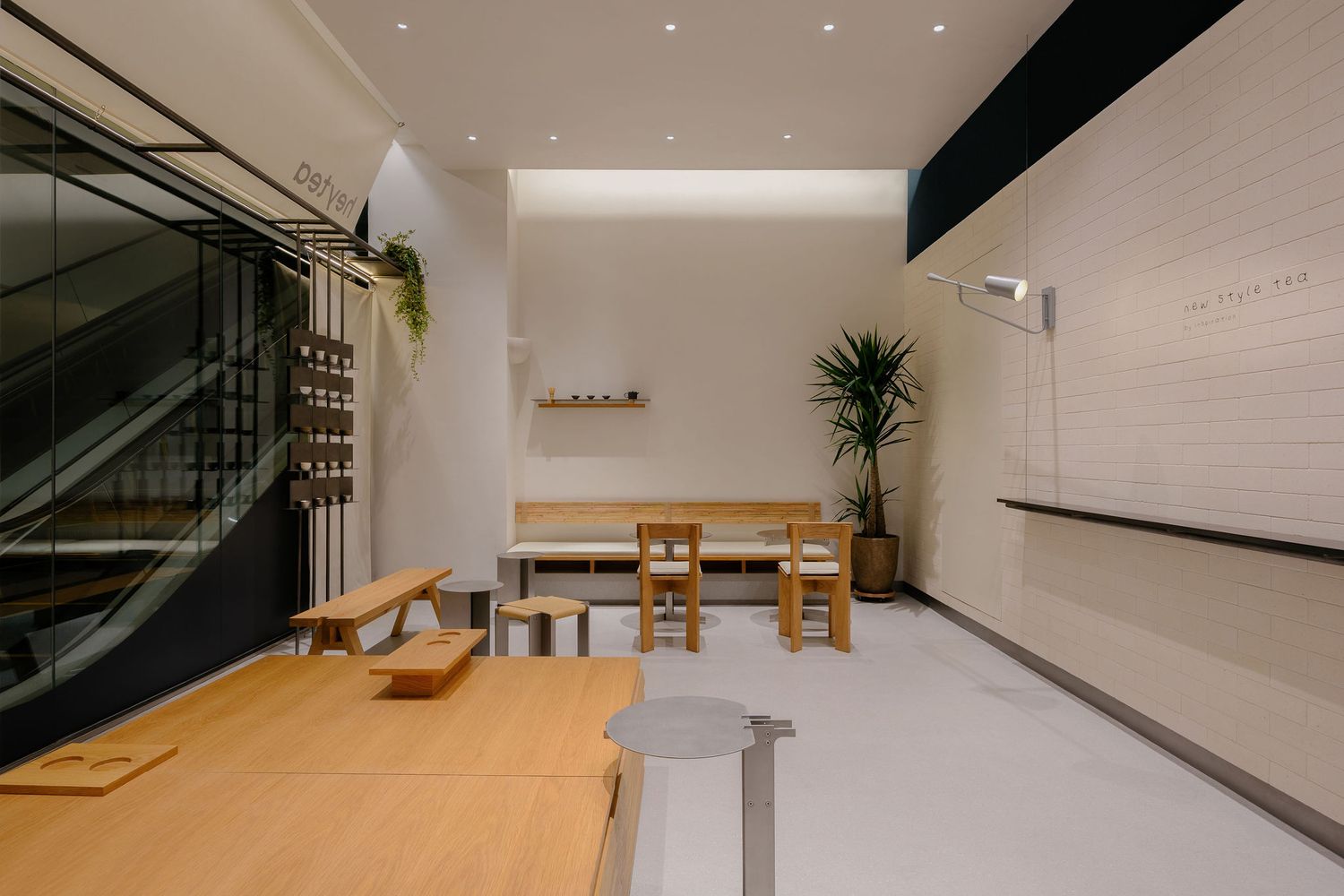 Minimalist shop interior with light wood benches, chairs and a round table, white walls, a potted plant and a glass staircase on the left.