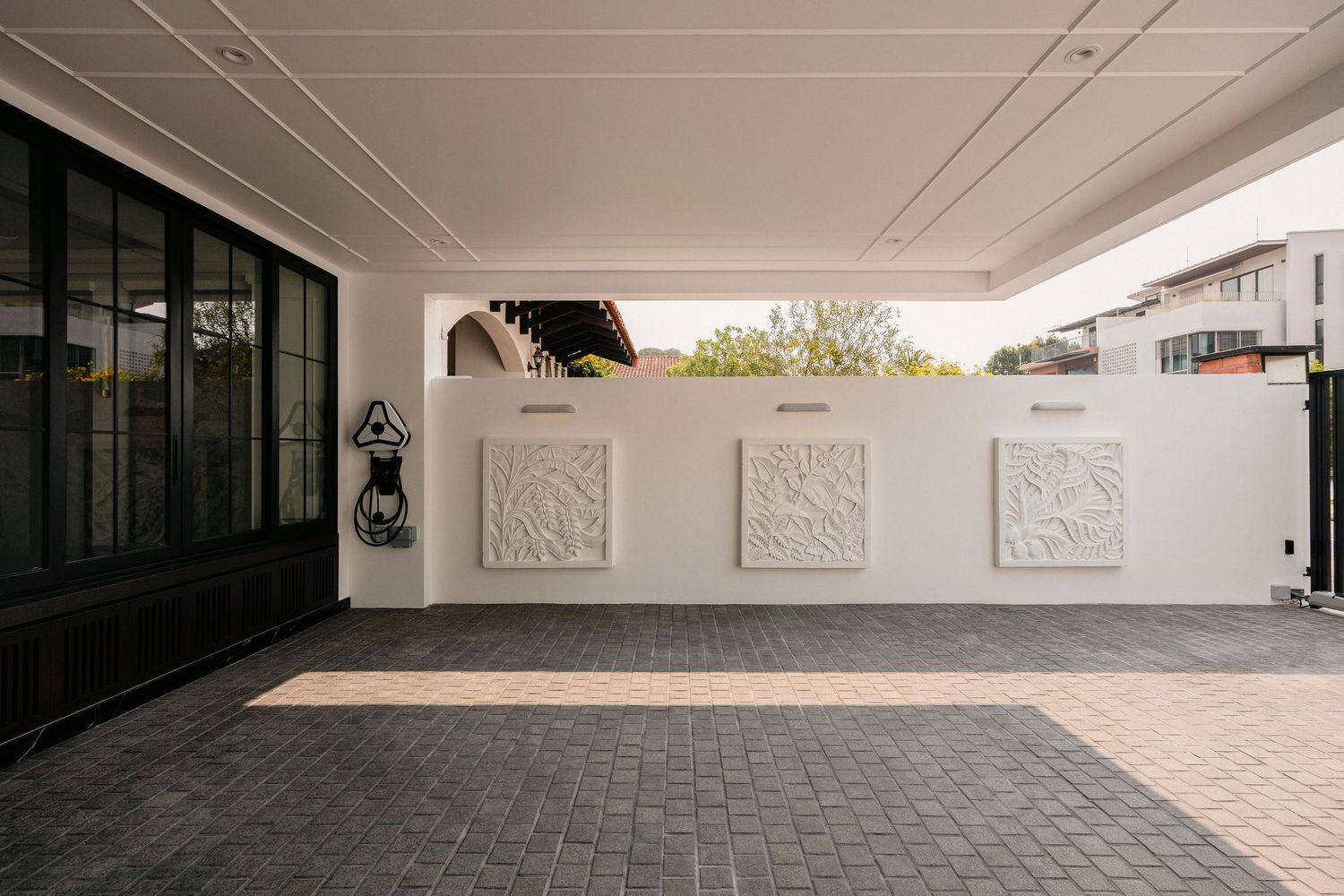 Covered carport with dark brick paving, a white feature wall with three carved decorative relief panels, and black framed glass doors on the