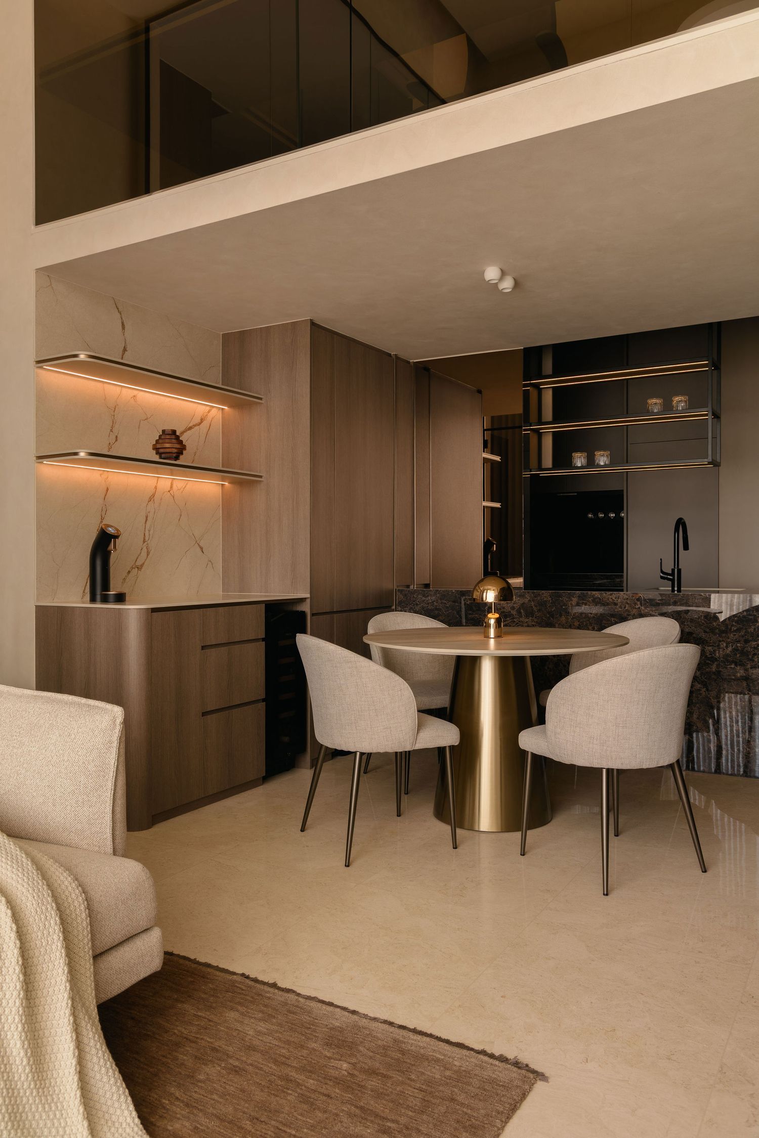Modern loft apartment with a round brass dining table, beige upholstered chairs, warm wood cabinetry and backlit marble shelves.