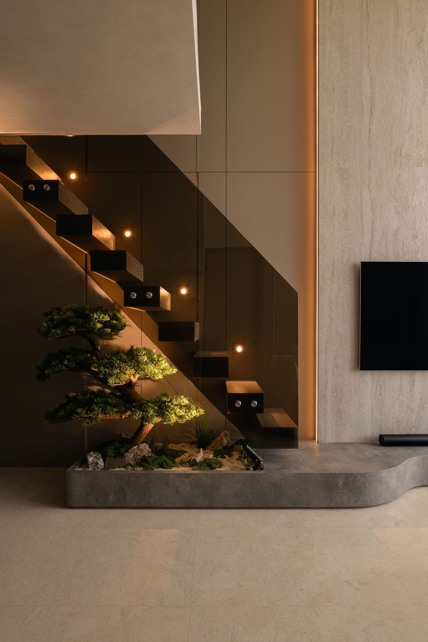Modern floating staircase with warm LED step lighting above an indoor bonsai tree garden bed, beside a travertine wall and wall-mounted TV.