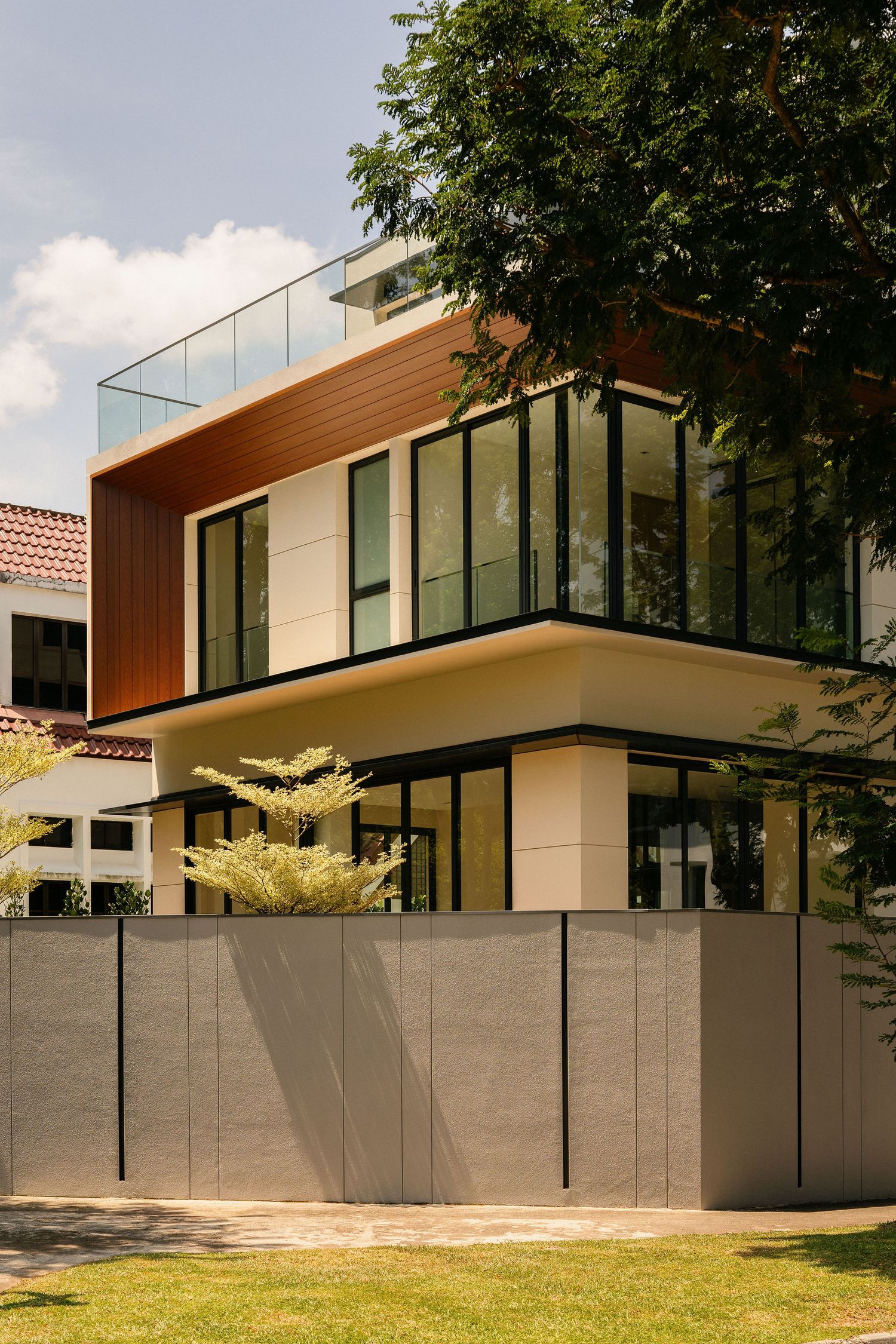 Modern two-story house with floor-to-ceiling glass windows, wood cladding, a glass balcony, and a gray perimeter wall beside a green lawn.