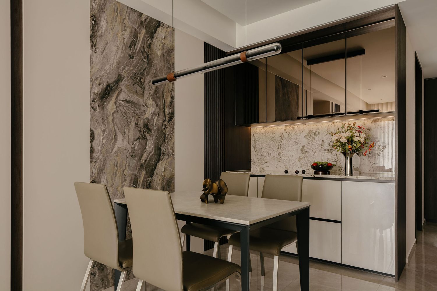 Modern dining area with a white table, beige chairs, marble accent wall, pendant light and a sleek built-in cabinet with fresh flowers.