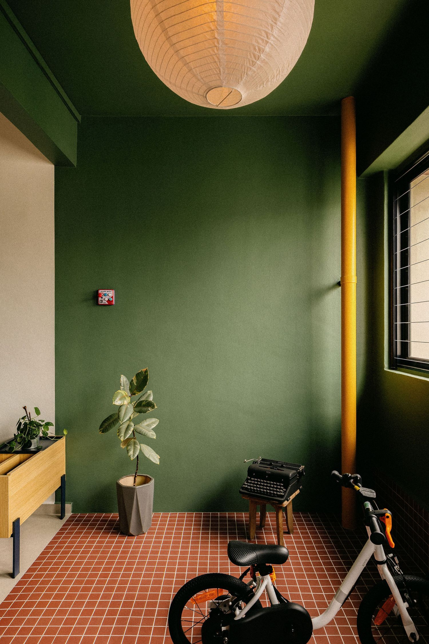Green-walled room with terracotta tile floor, yellow pipe, paper lantern light, potted plant, vintage typewriter and a white kids bike.