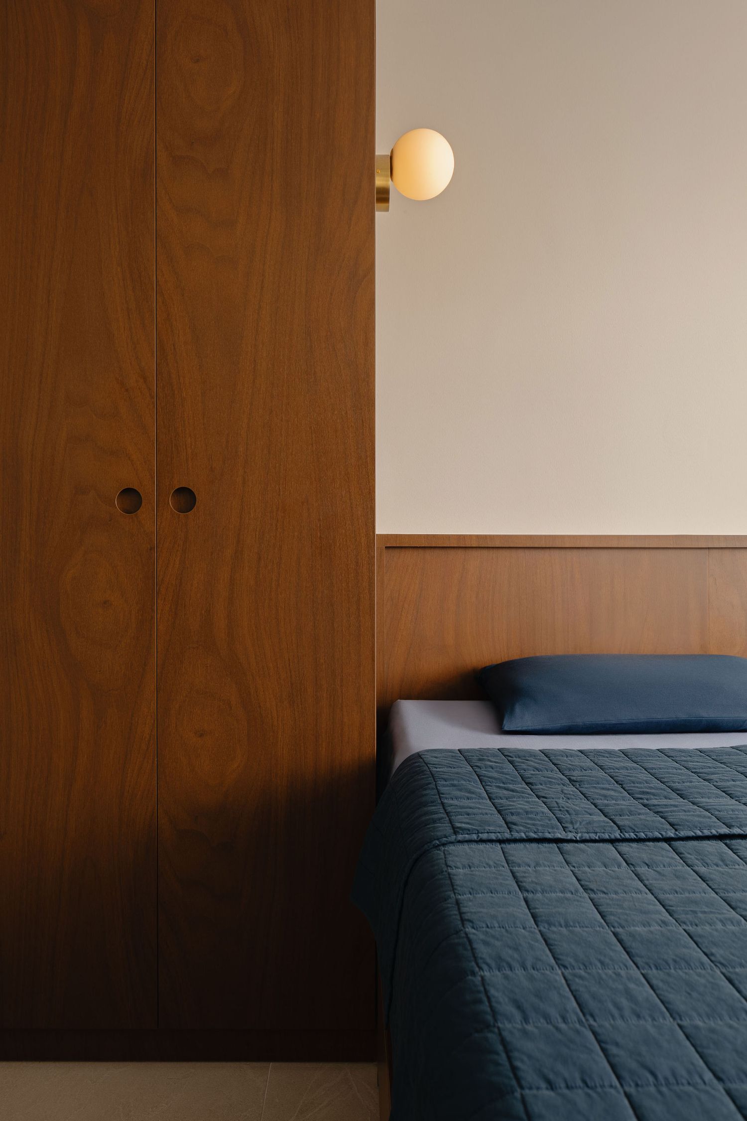 Mid-century modern bedroom with walnut wardrobe, blue quilted bedspread, wood headboard panel, and gold globe wall sconce.