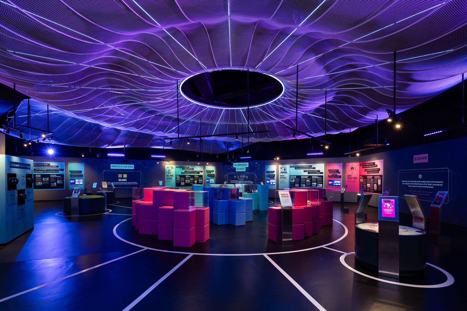 Futuristic museum exhibit hall with glowing purple neon ceiling lights, colorful interactive displays, and circular floor patterns.