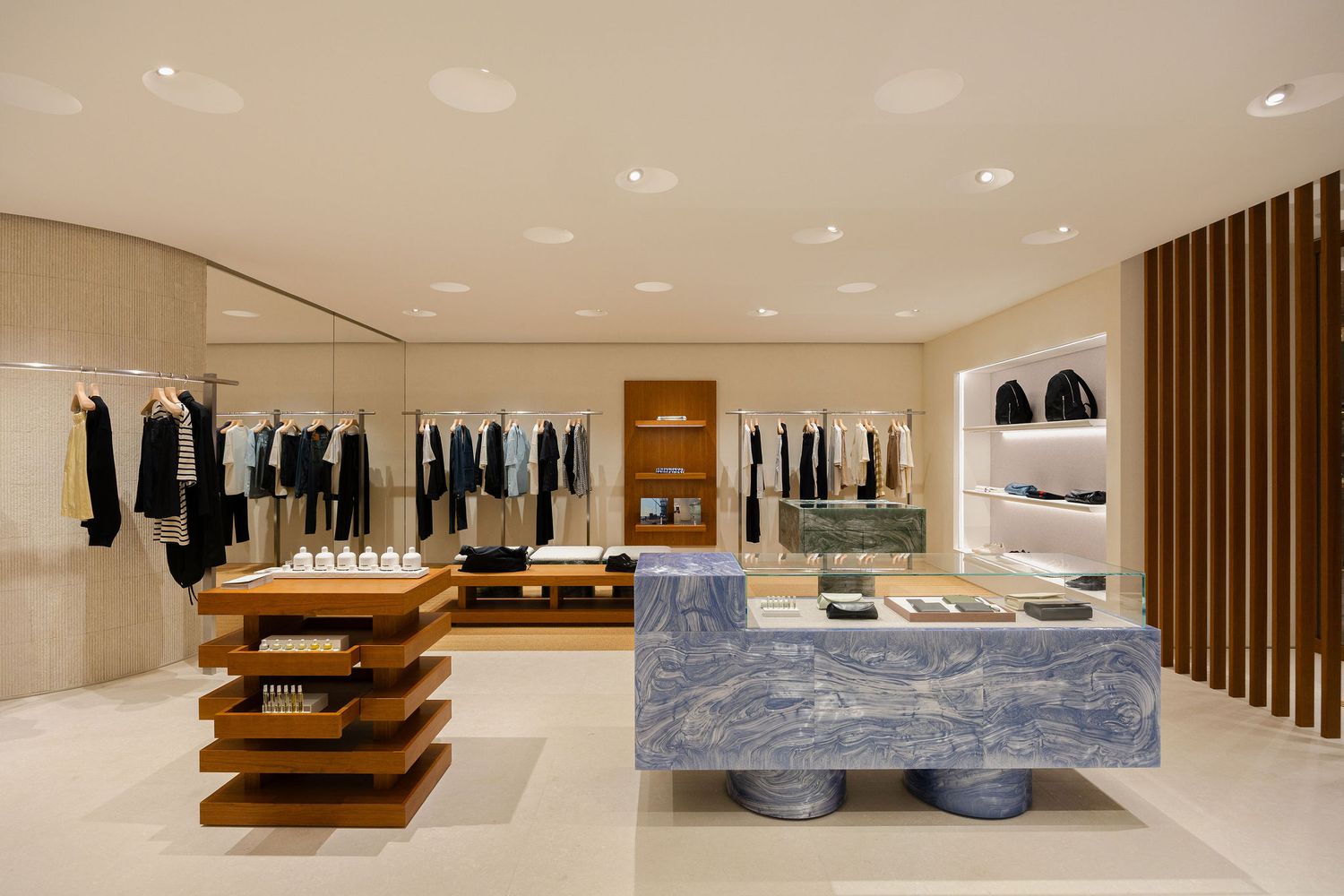Modern luxury retail store interior with wooden display tables, marble counter, and clothing racks in a minimalist beige space.