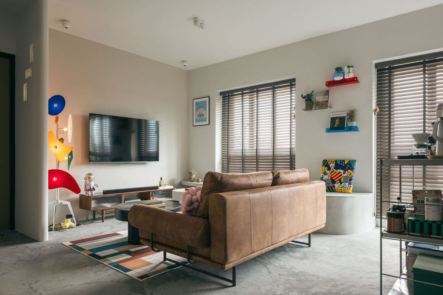 Modern living room with brown leather sofa, wall-mounted TV, wooden blinds, and colorful eclectic decor accents.