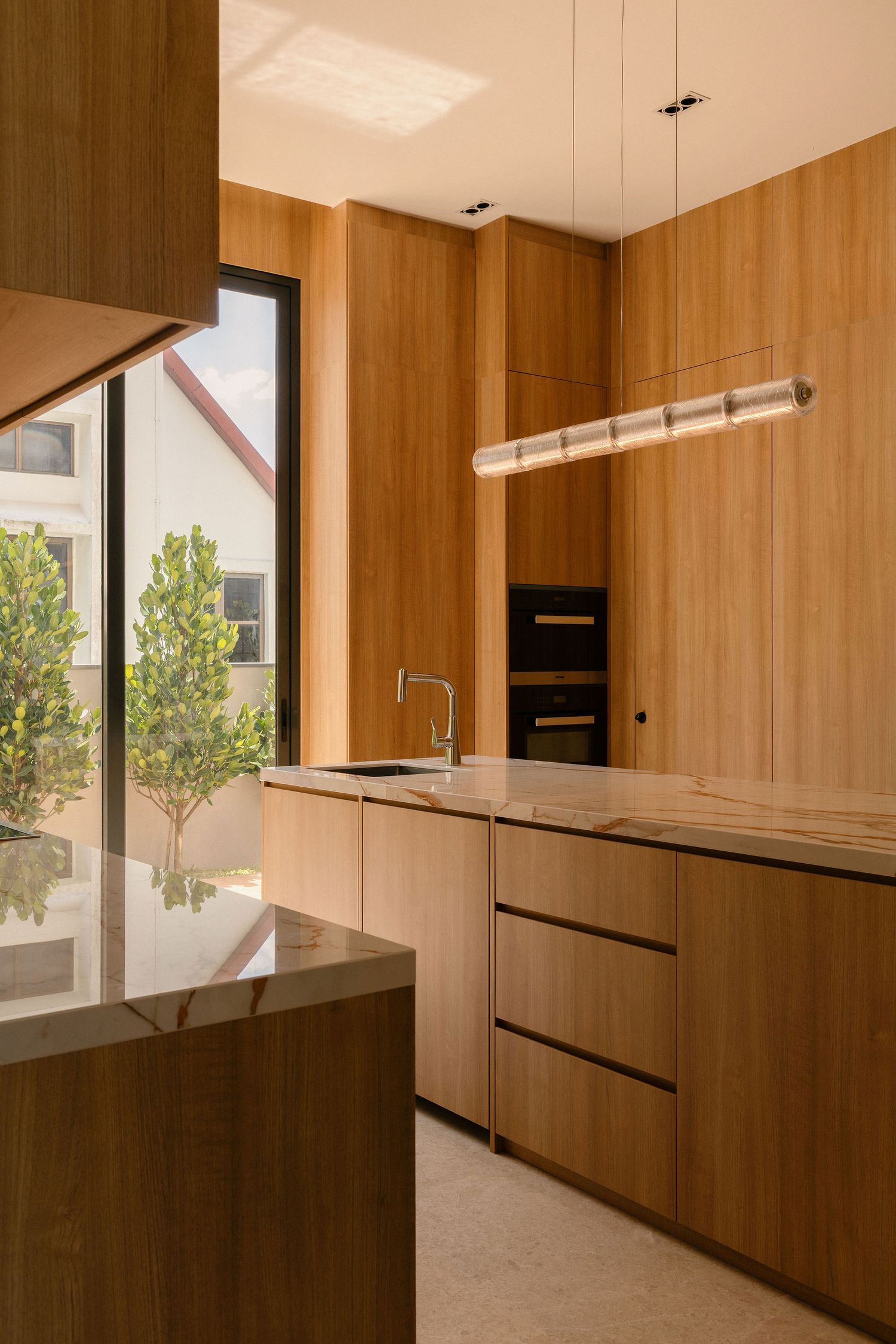 Modern kitchen with warm wood cabinetry, marble countertops, built-in ovens, a linear pendant light and a large window overlooking trees.