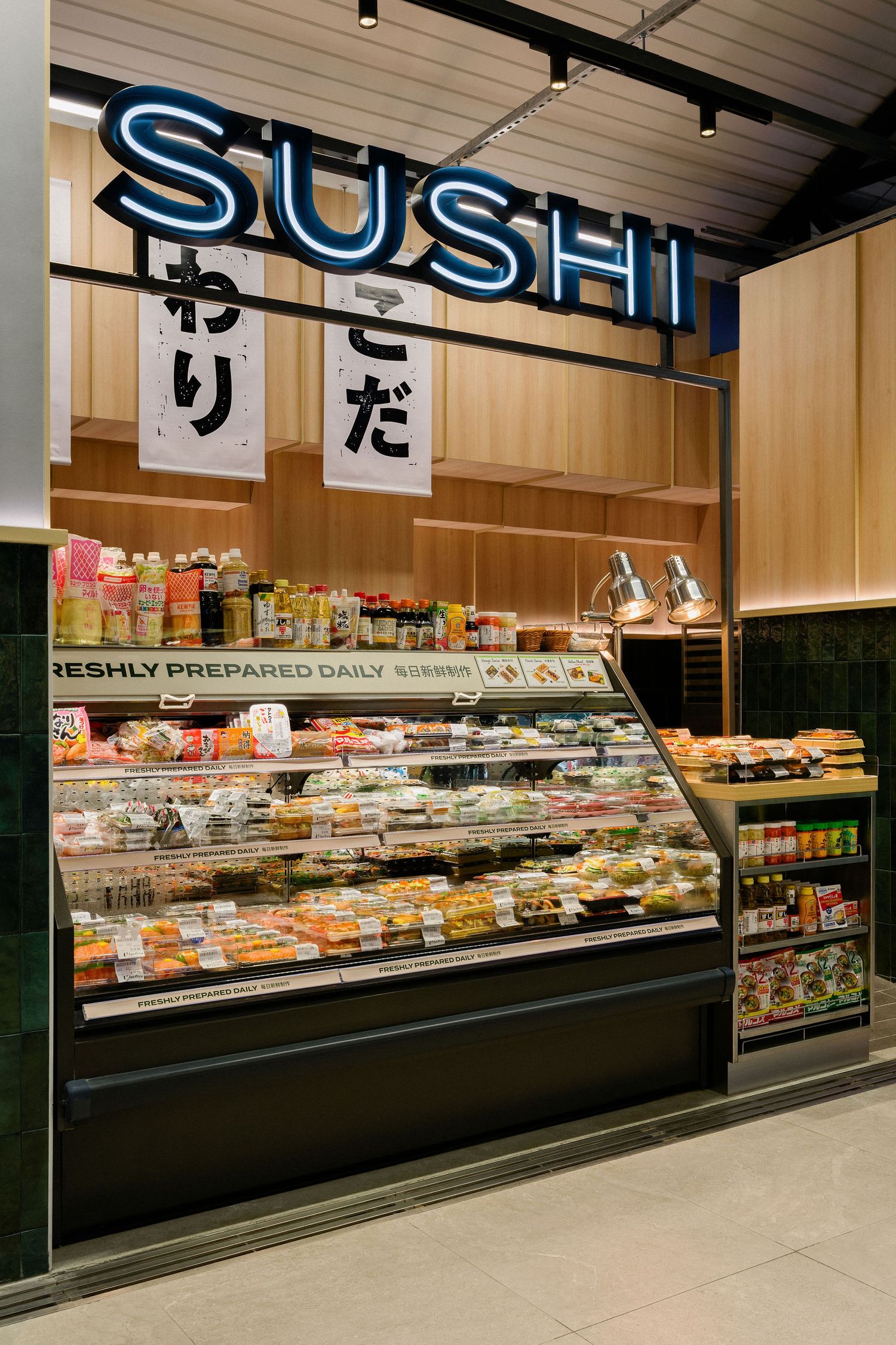 Neon SUSHI sign above a refrigerated display case of freshly prepared daily sushi packs in a modern Japanese grab-and-go shop.