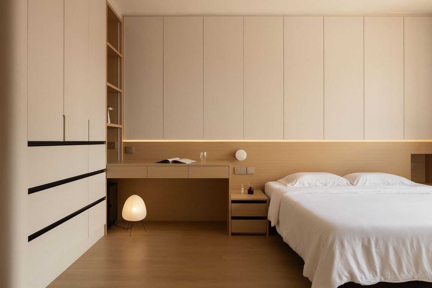 Minimalist bedroom with built-in wardrobe, wooden desk, white bedding, and warm neutral tones.