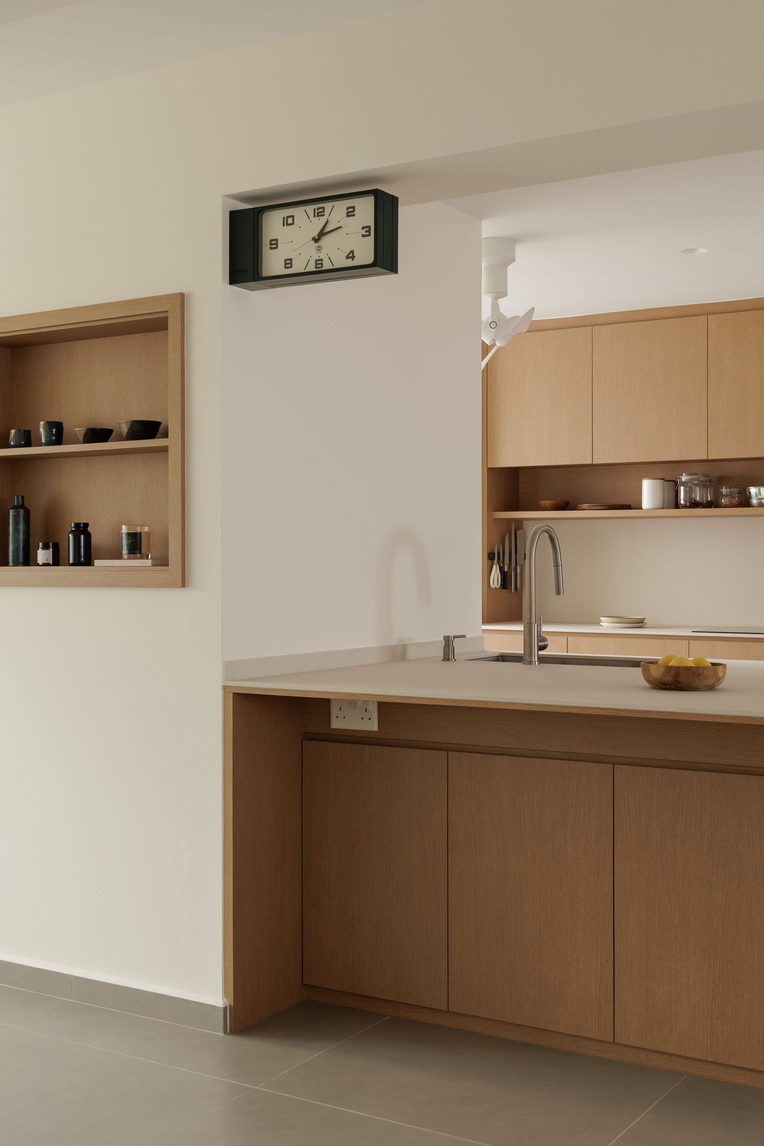 Modern minimalist kitchen with light wood cabinets, built-in shelving, and a wall-mounted clock above the counter space.