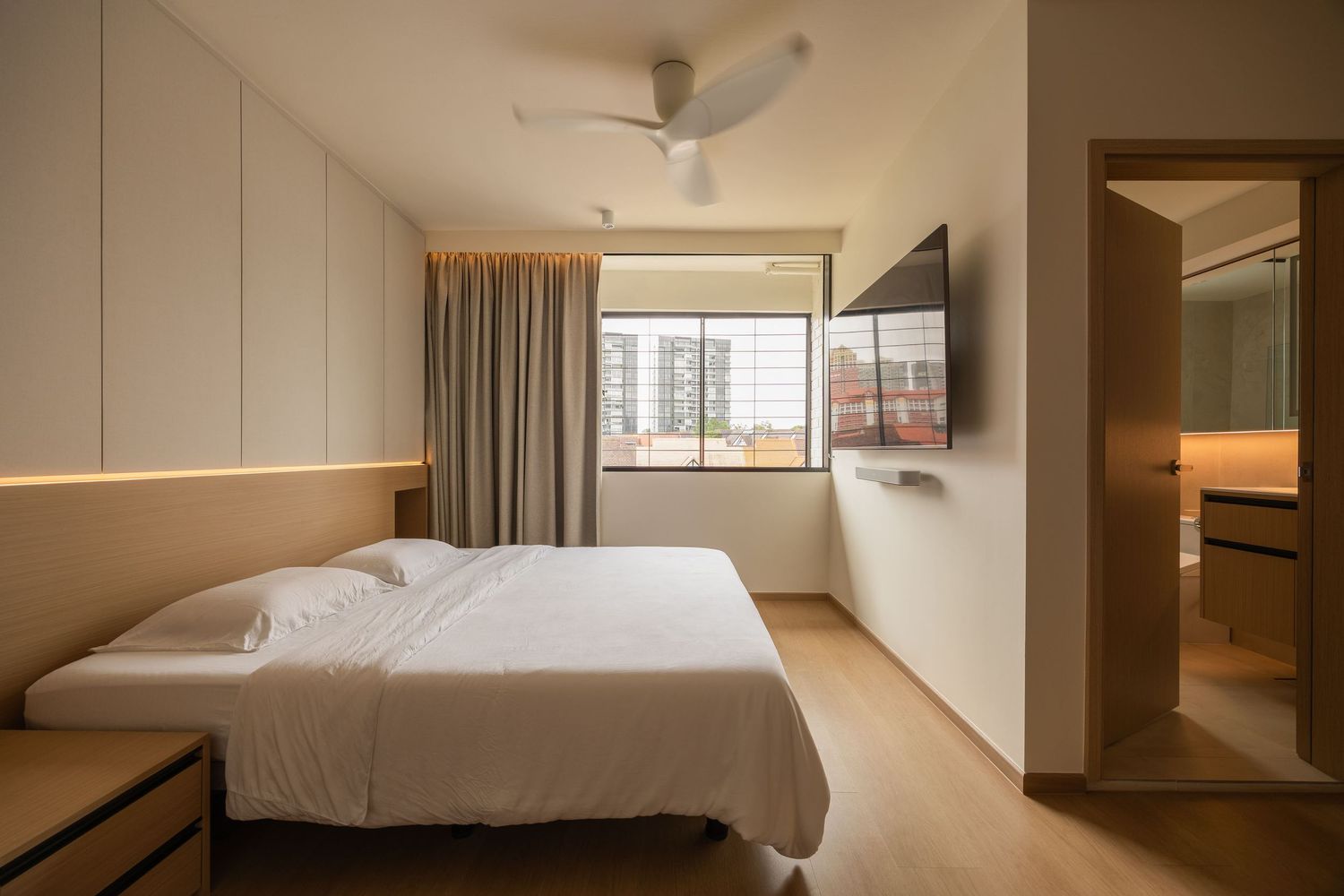 Modern hotel room interior with white bed, wall-mounted TV, ceiling fan, and attached bathroom visible through doorway.