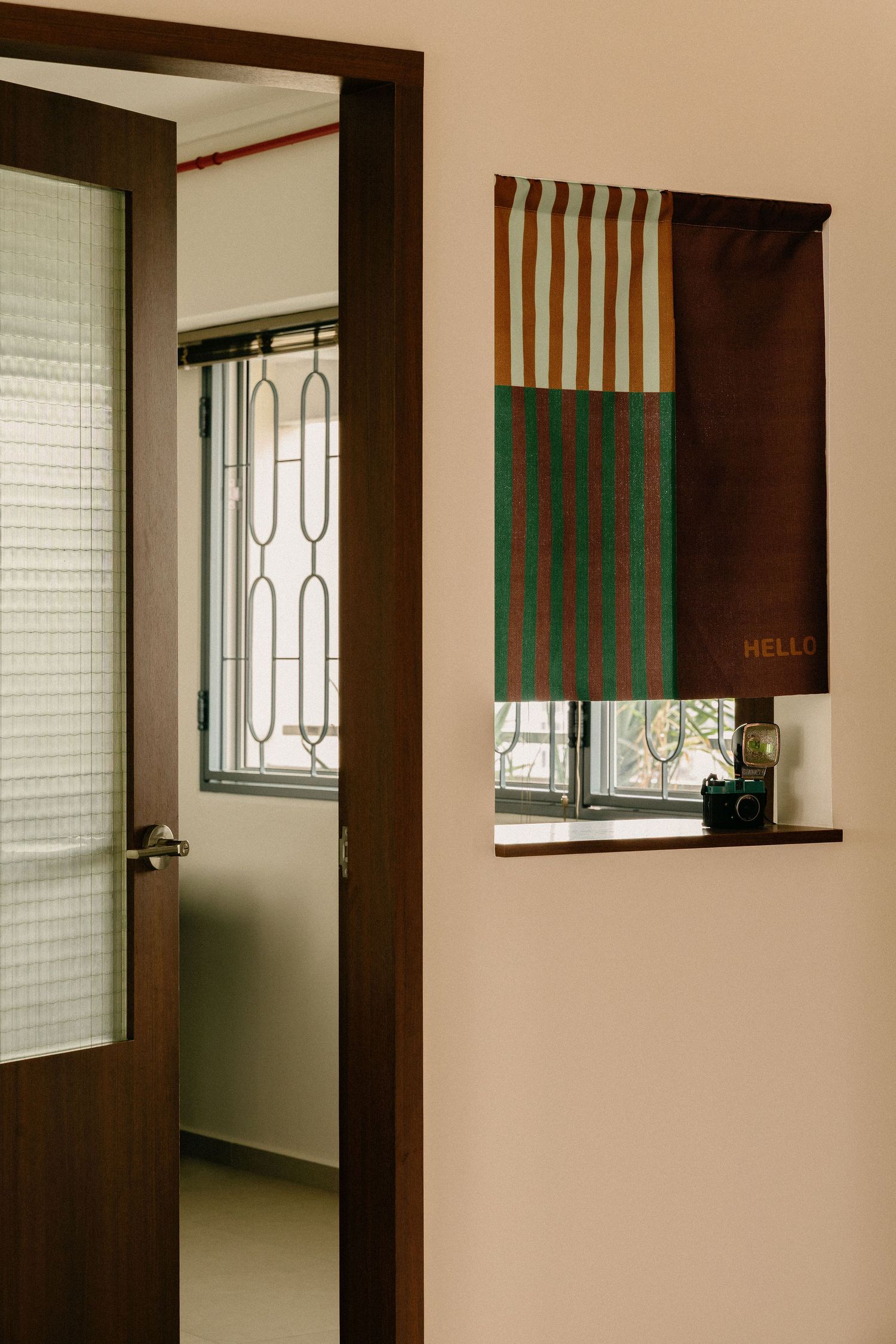 Open brown wooden door beside a window with a striped green, brown and cream fabric blind that reads HELLO, with a camera on the sill.