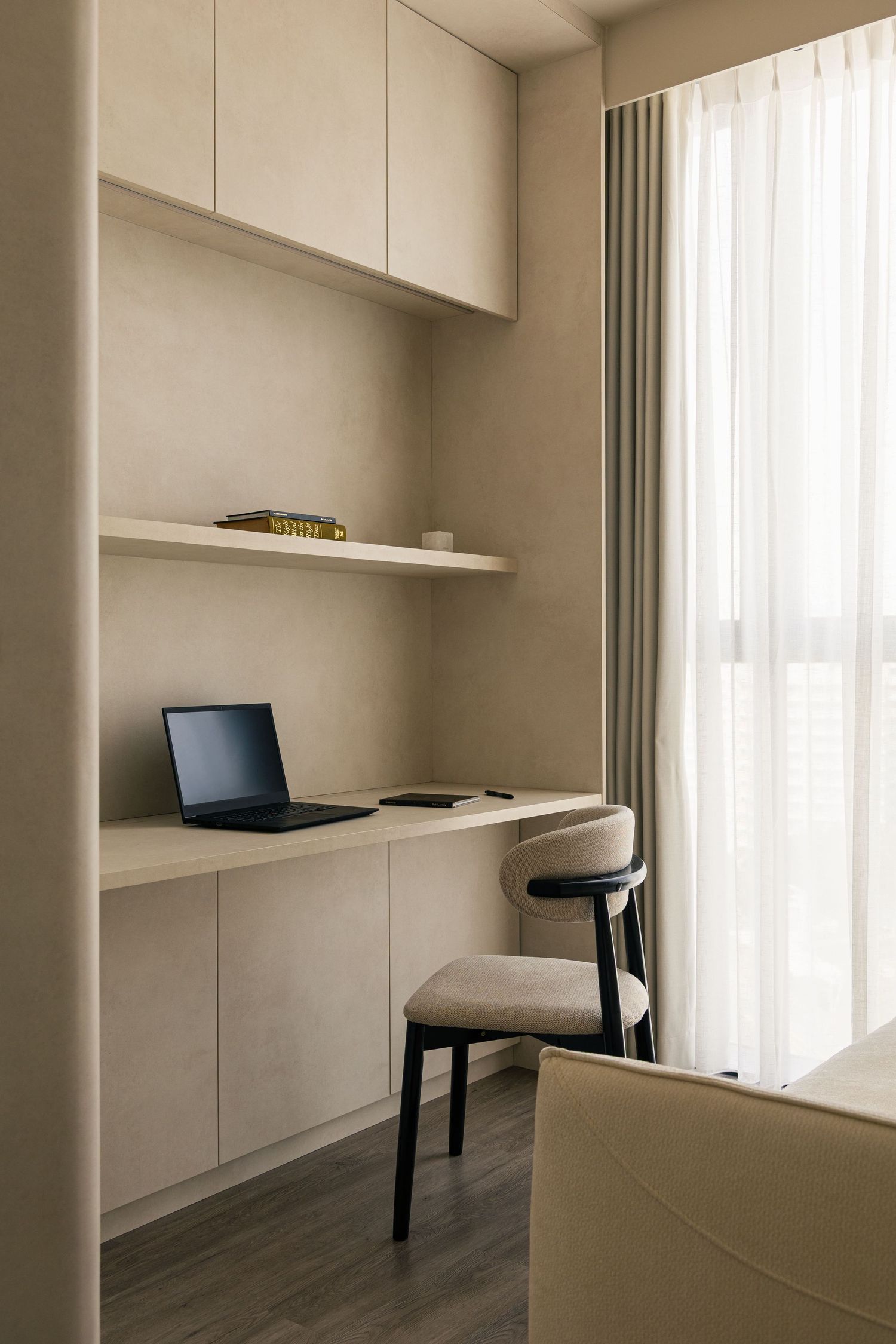 Minimalist built-in desk with laptop, overhead cabinets, open shelf, and neutral tones beside a sheer curtained window.