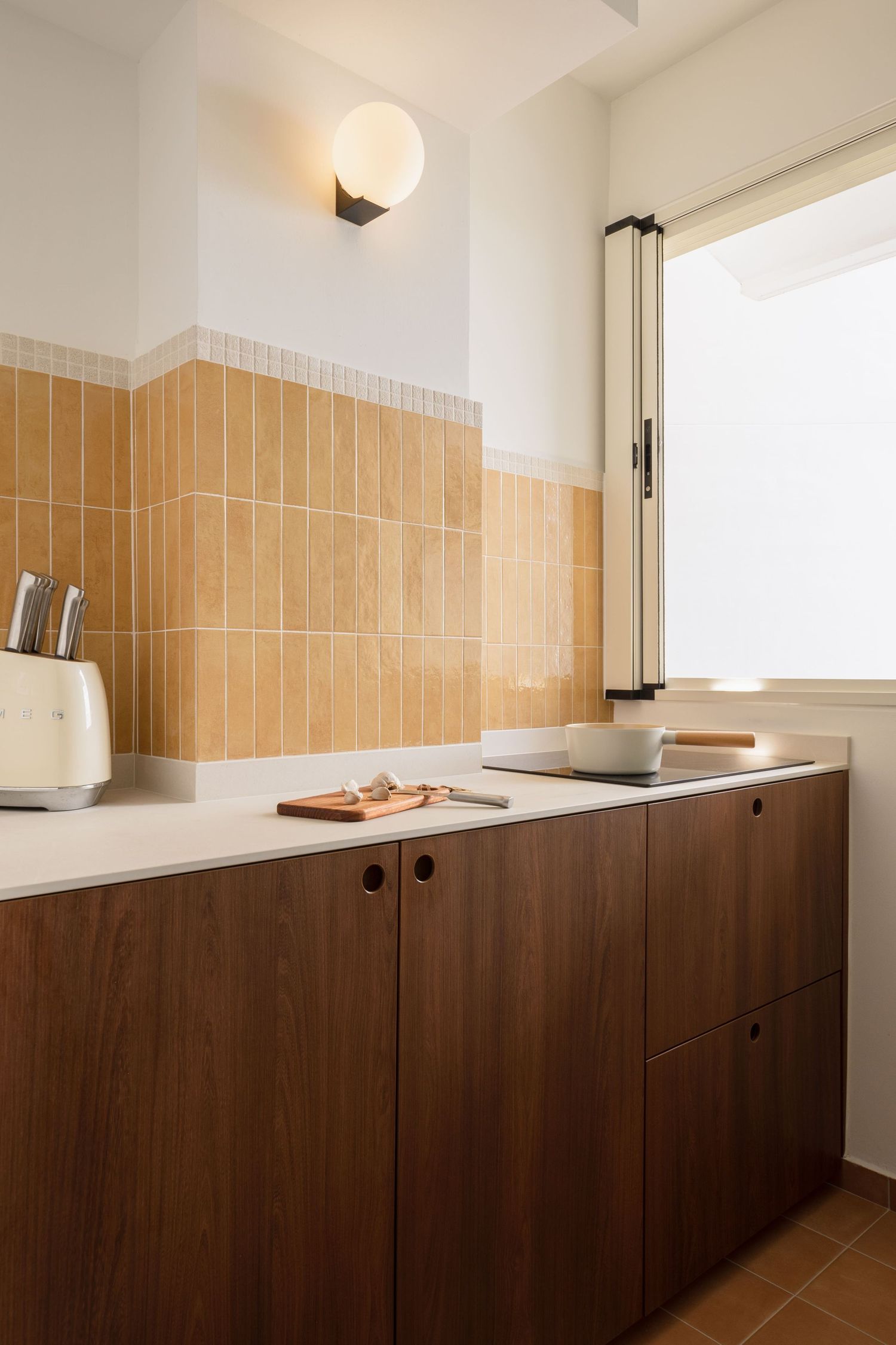 Modern kitchen with warm terracotta tiles, dark walnut cabinets, white countertop, toaster, and globe wall sconce near window.