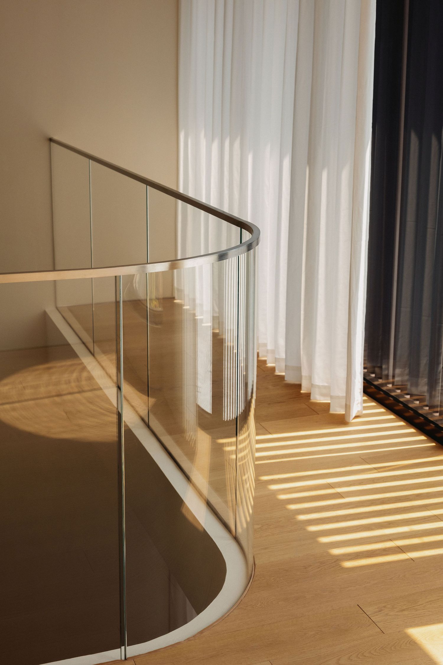 Modern curved glass staircase railing with metal handrail on hardwood floor, bathed in natural sunlight through sheer curtains.