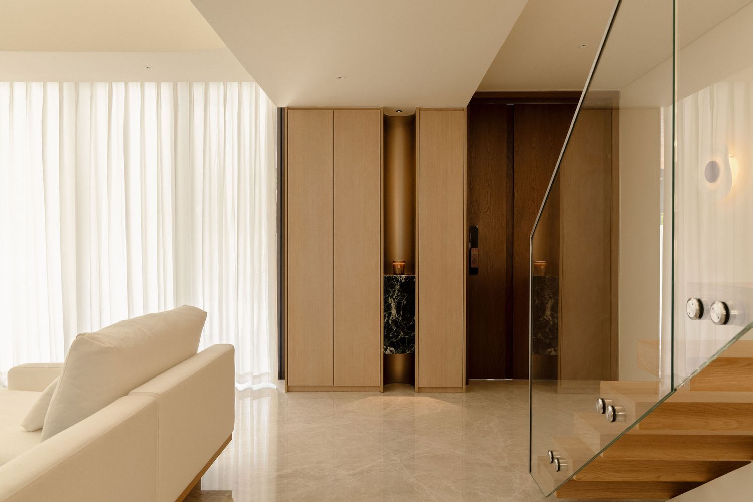 Modern minimalist living room with white sofa, glass staircase railing, and warm wood cabinetry against neutral walls.