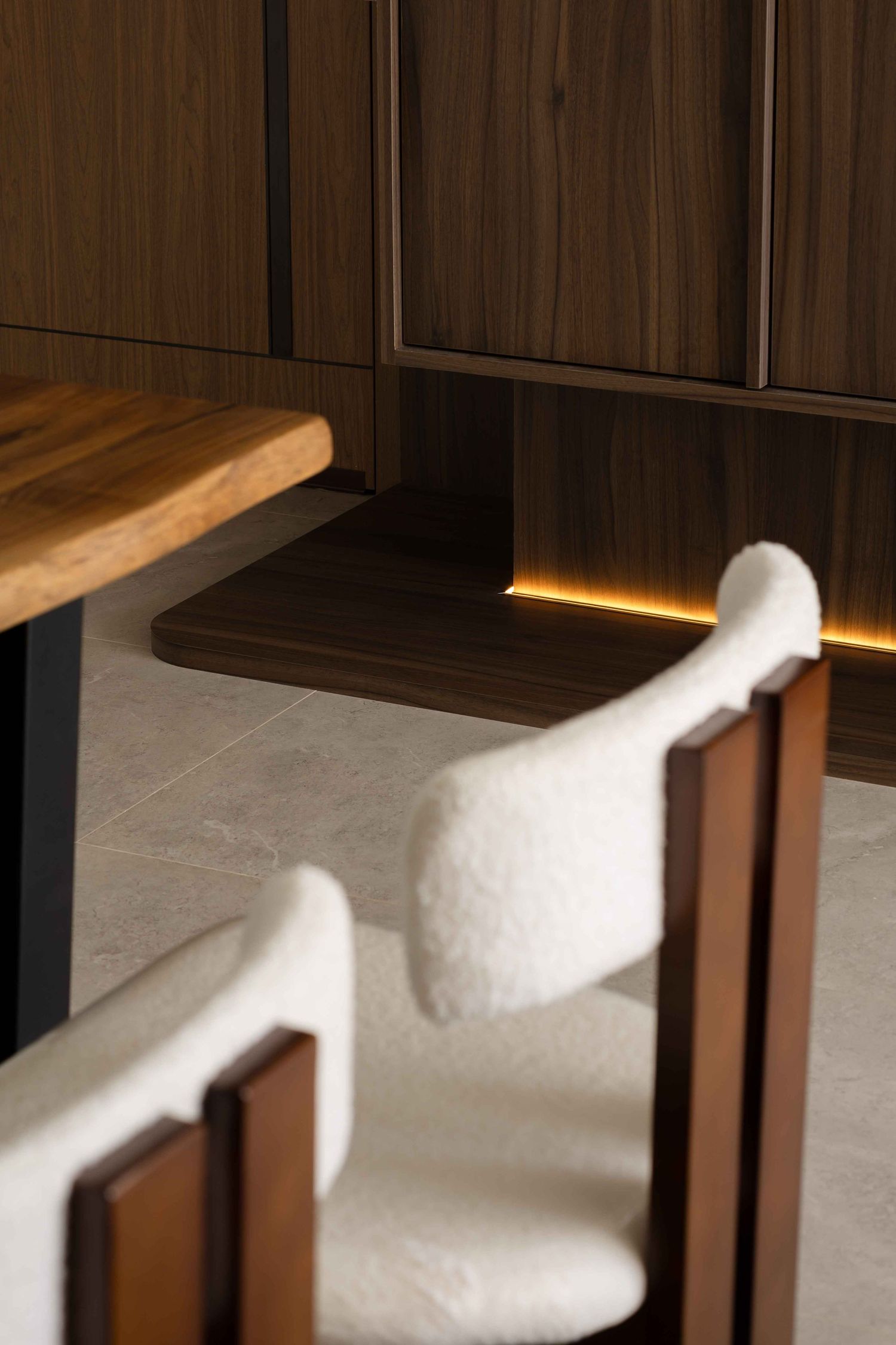 Modern dining area with white furry chairs and wooden table against dark wood paneled wall with ambient lighting.